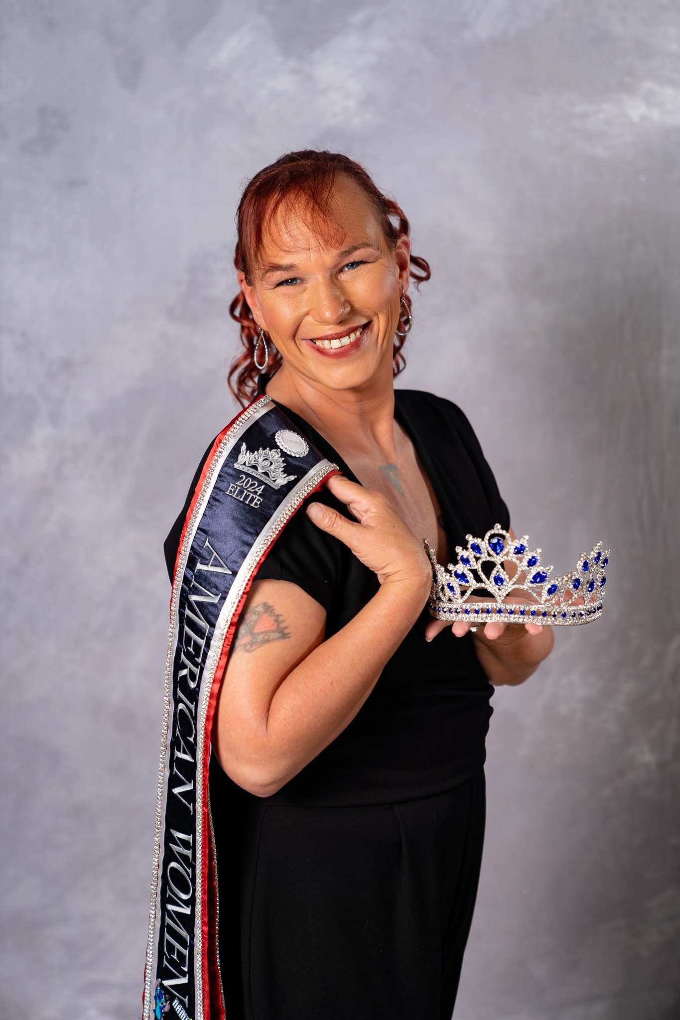 Smiling woman holding jeweled pageant crown while wearing American Women sash in black outfit.