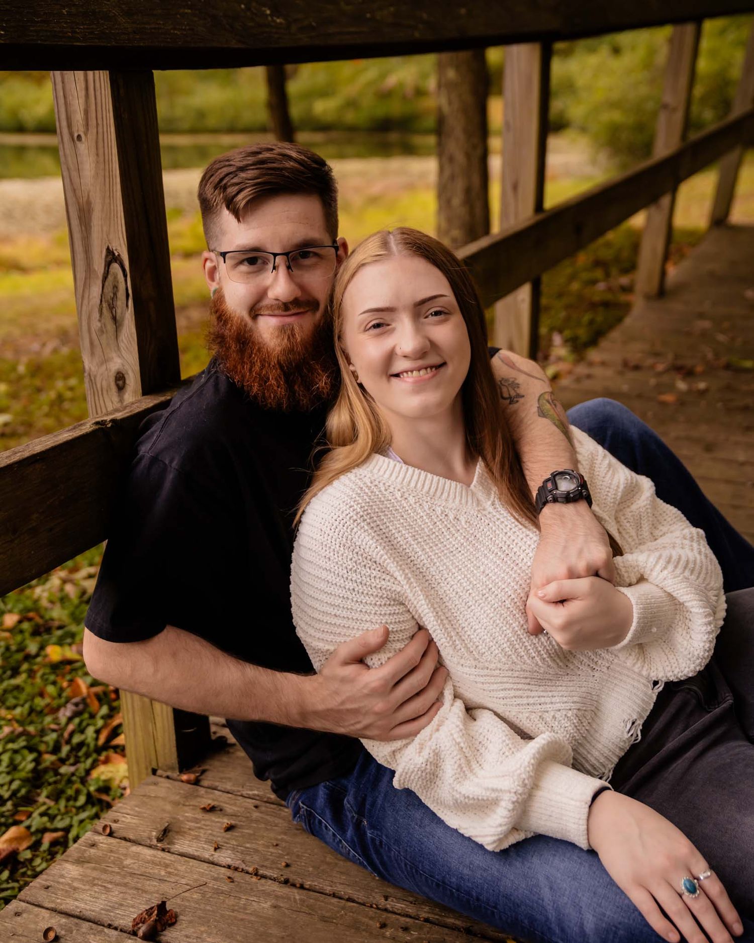 A smiling couple sits together on a wooden bridge deck, embracing in a cozy autumn outdoor setting.