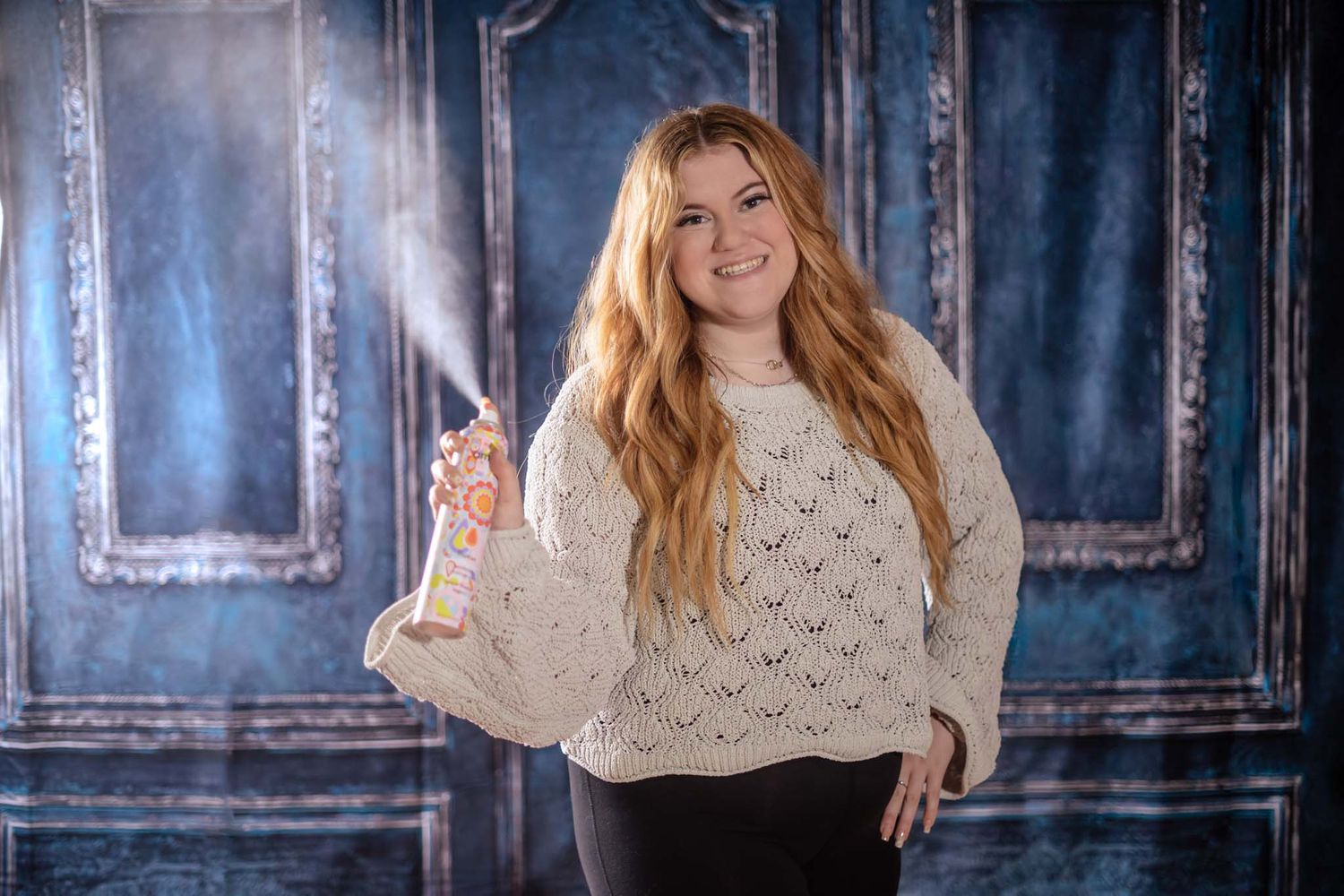 Smiling young woman in cream knit sweater holding spray bottle, blue door backdrop.