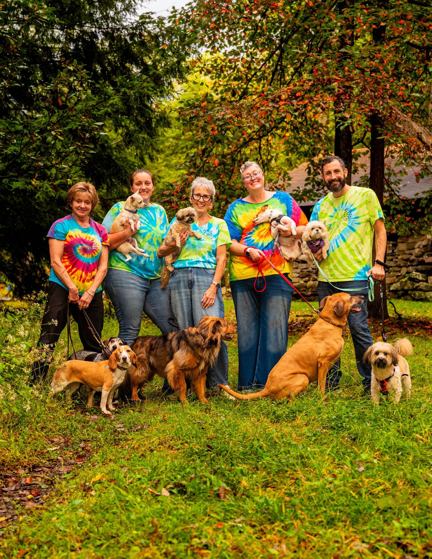 Group of five people in colorful tie-dye shirts posing with multiple dogs on green grass in autumn.