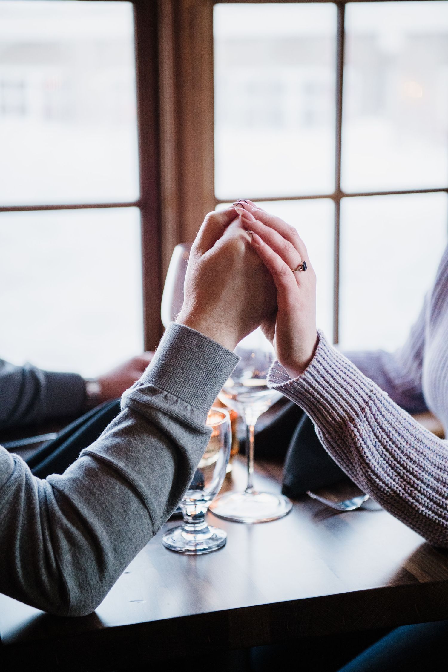 Couple holding hands tenderly over a dark wooden table with wine glasses, sitting by a bright window in a cozy restaurant.