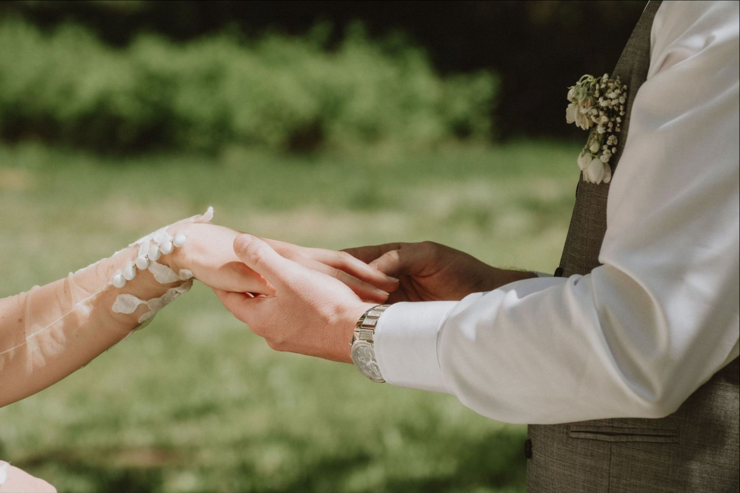 Interracial couple exchanging wedding rings outdoors, groom wearing gray vest with boutonniere during ceremony.