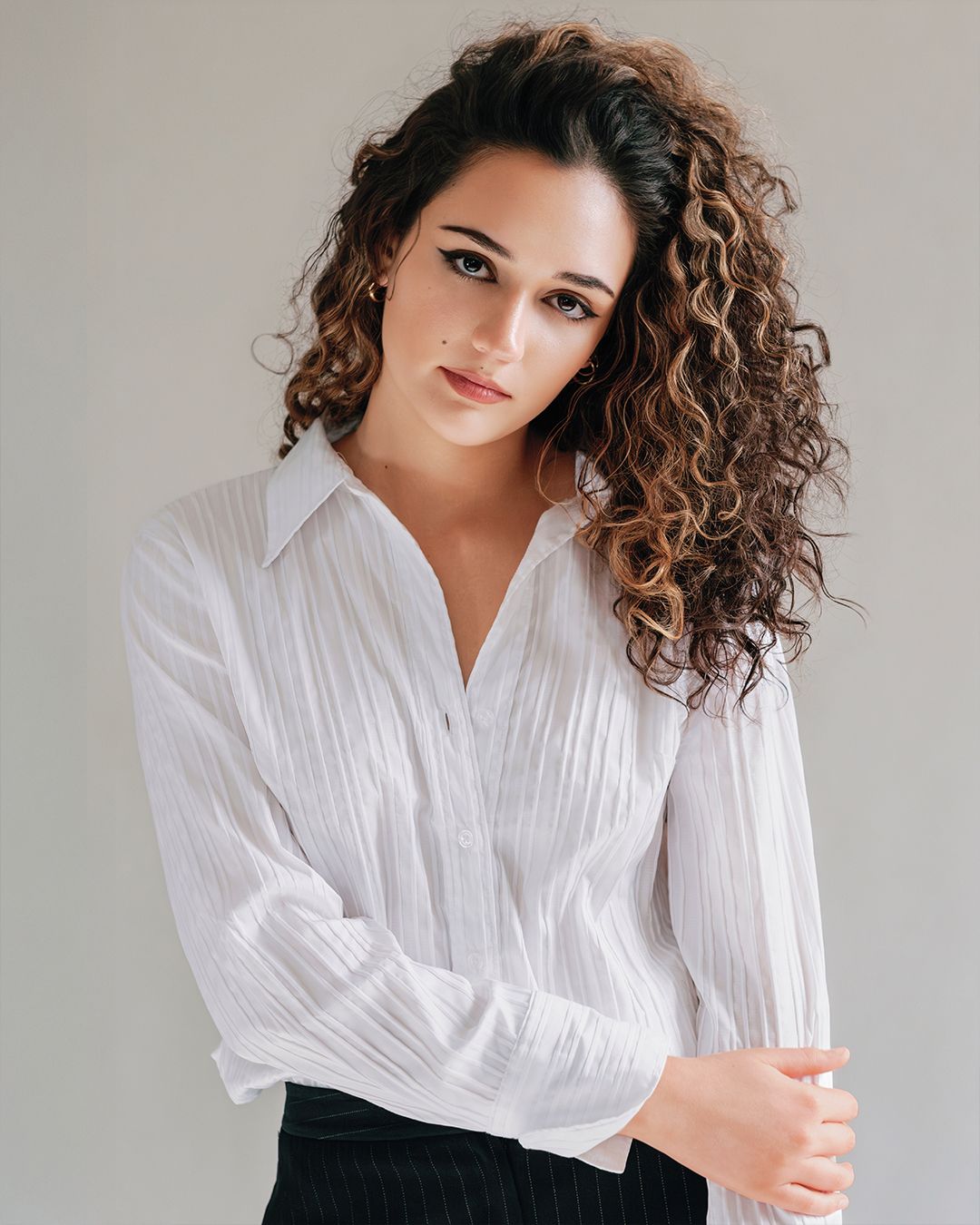 Young woman with curly brown hair wearing a white button-up shirt poses against a neutral background.