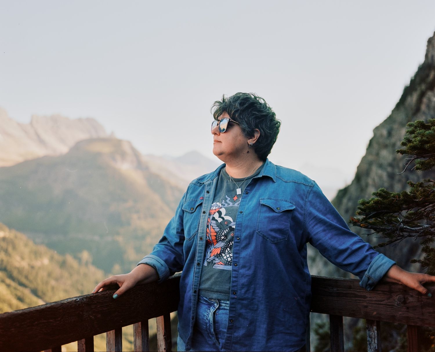 A person with blue hair and sunglasses enjoying a mountain overlook view.