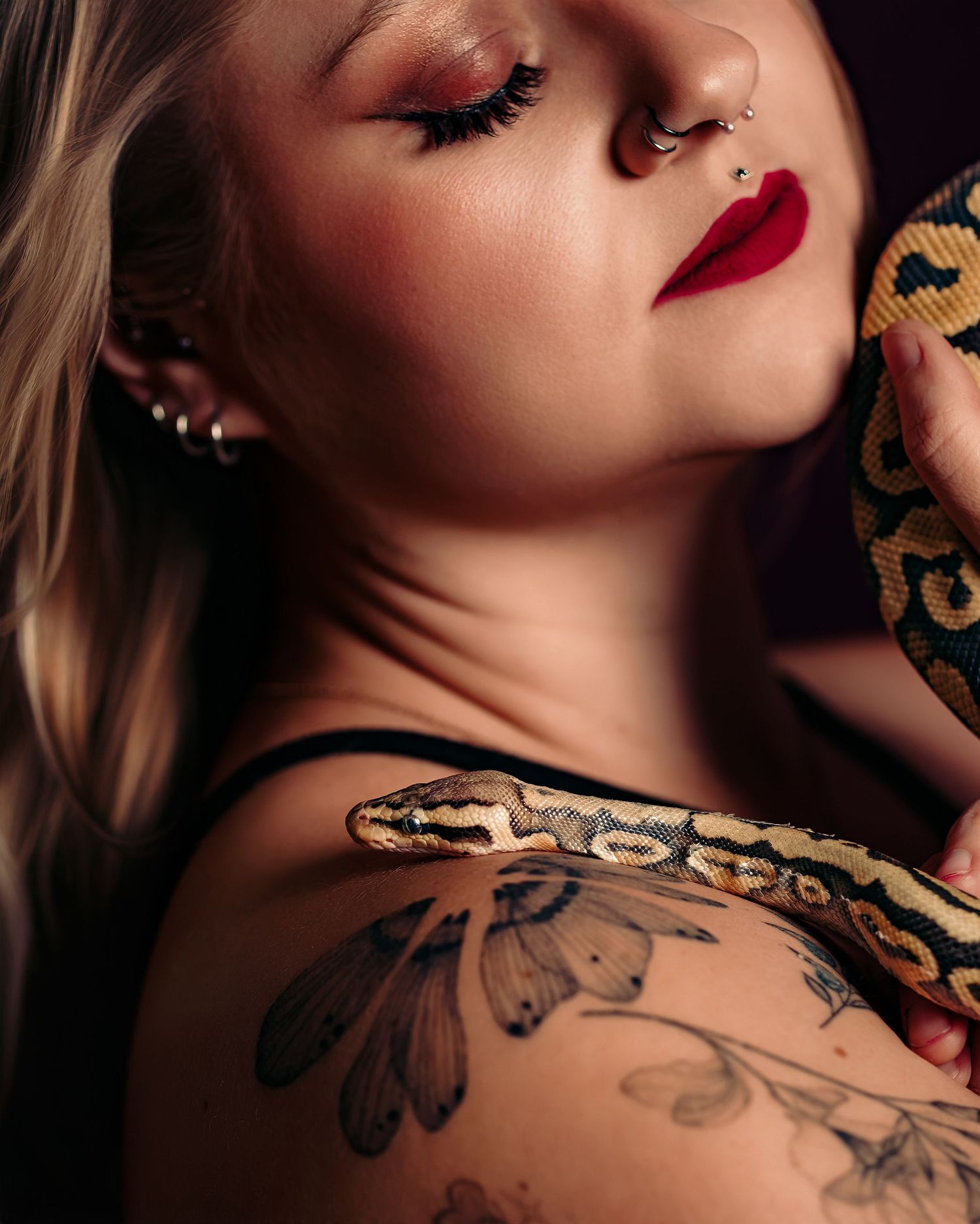 Woman with red lips, septum piercing, and floral tattoo poses with a ball python snake draped over her shoulder.