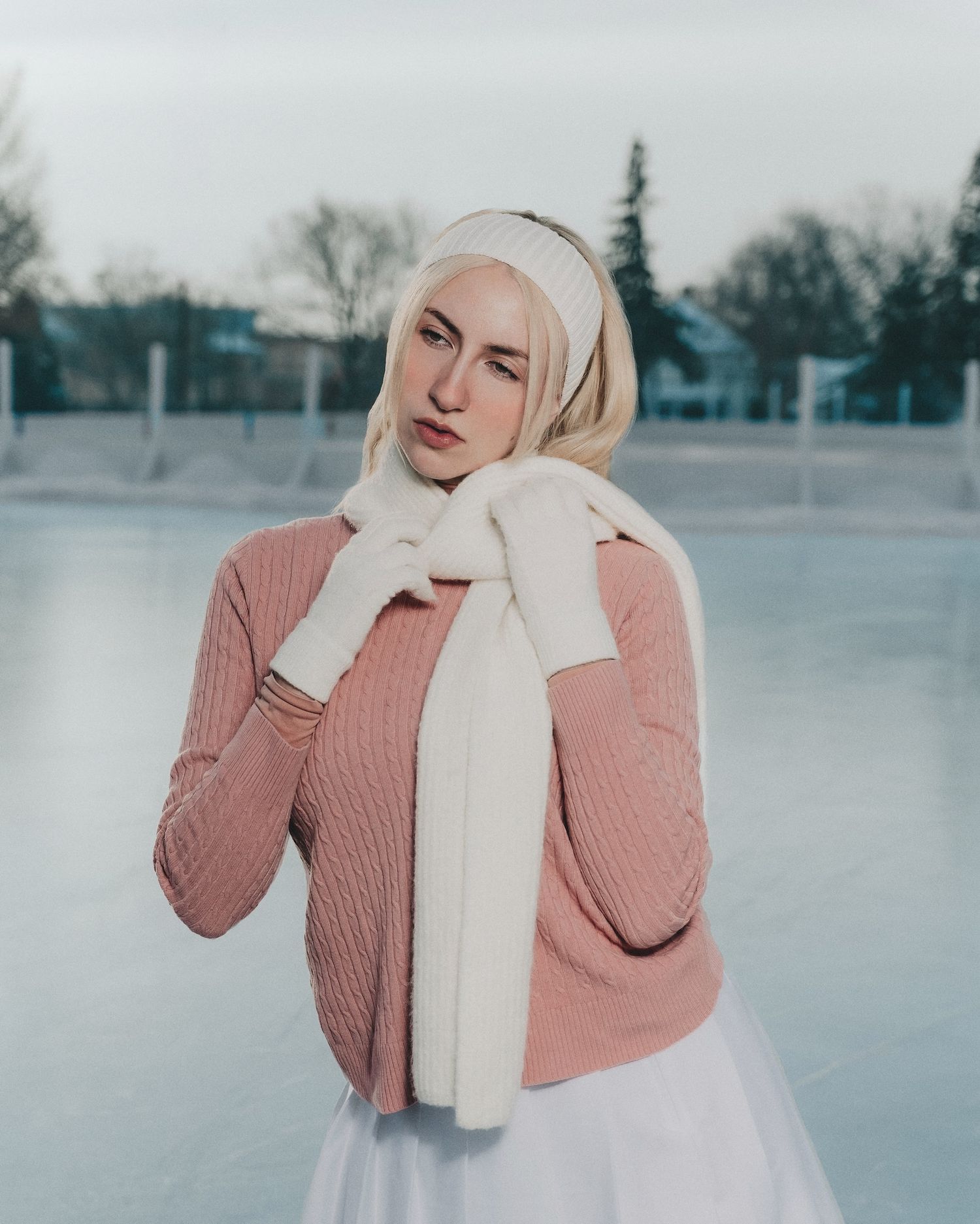 A blonde woman in a pink ribbed sweater, white scarf, and headband poses at an outdoor ice rink.