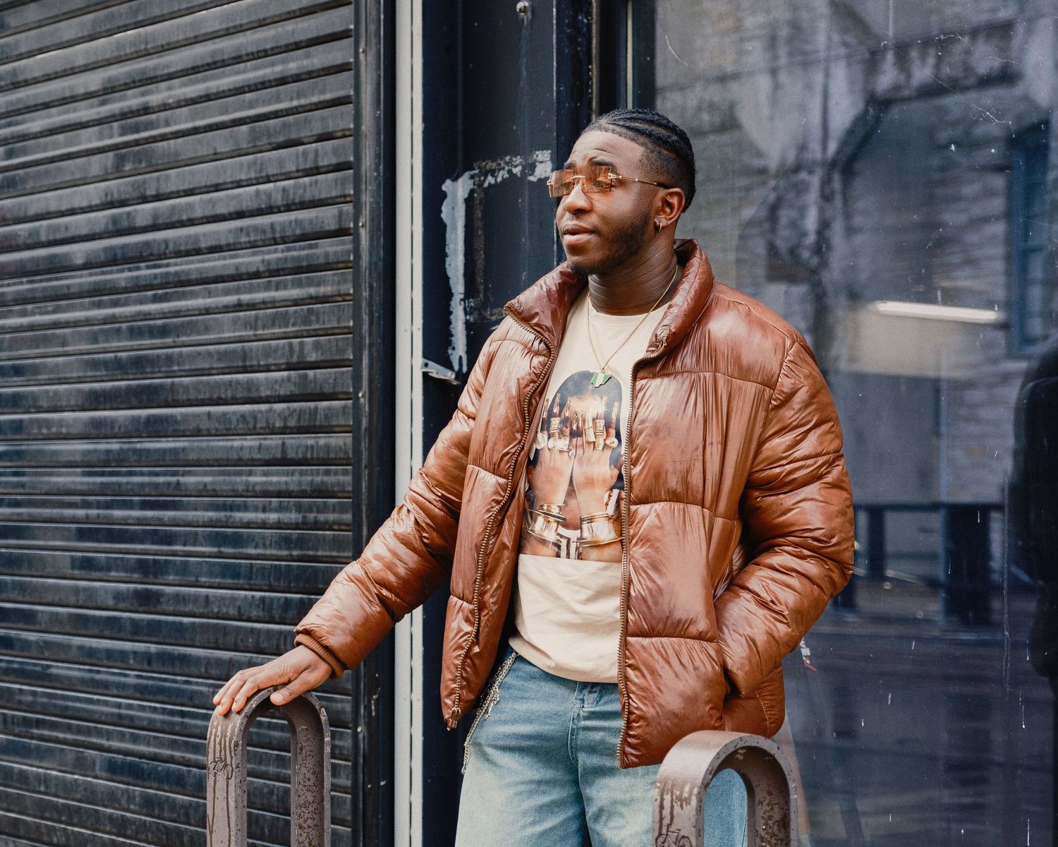 A man in a brown puffer jacket and jeans leans against a metal railing on a city street.