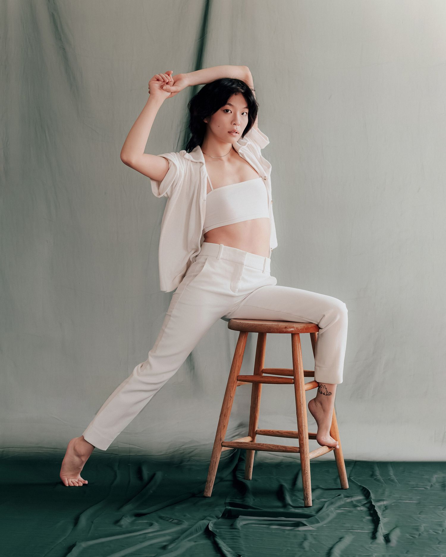 Woman in white crop top and pants poses gracefully on a wooden stool against a draped backdrop.