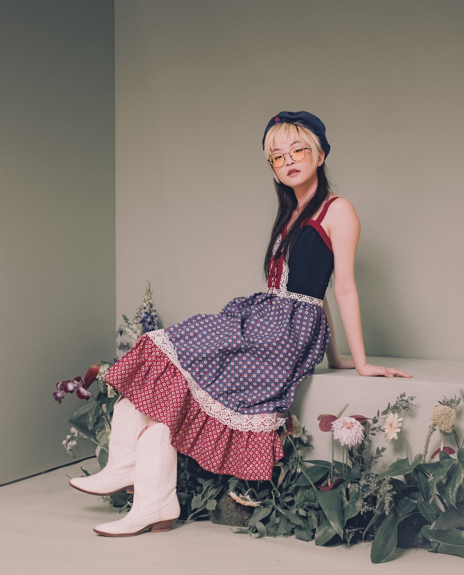 A young woman in a polka dot ruffled skirt and white cowboy boots sits among flowers in a styled studio setting.