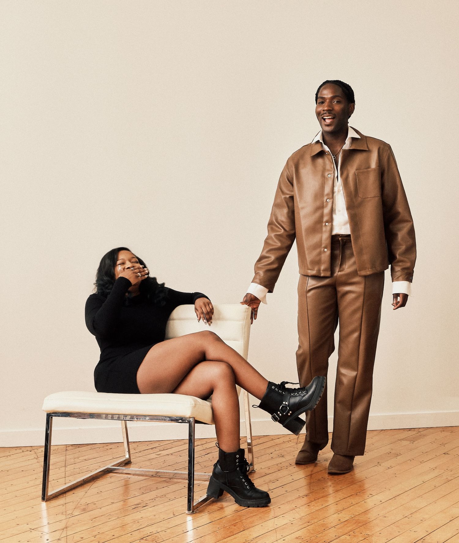 Two stylish people pose together, one seated in a chair and one standing, both wearing brown and black outfits.