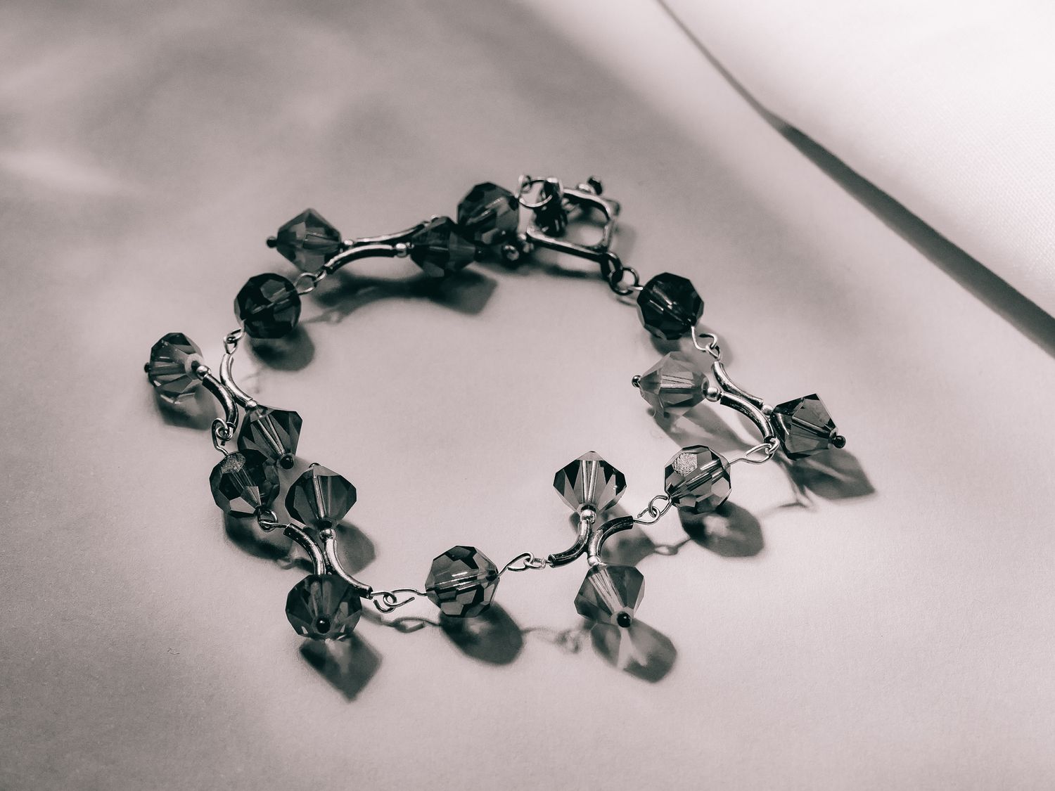 Monochrome close-up of a delicate crystal bead bracelet arranged in a circle on soft white fabric.