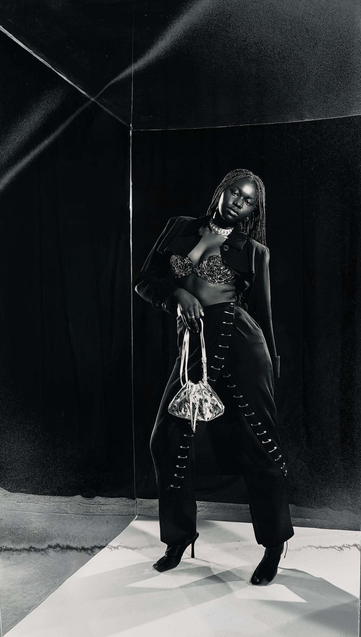 A black and white fashion photo of a woman in a blazer, lace bralette, wide-leg pants, and heels holding a bag.