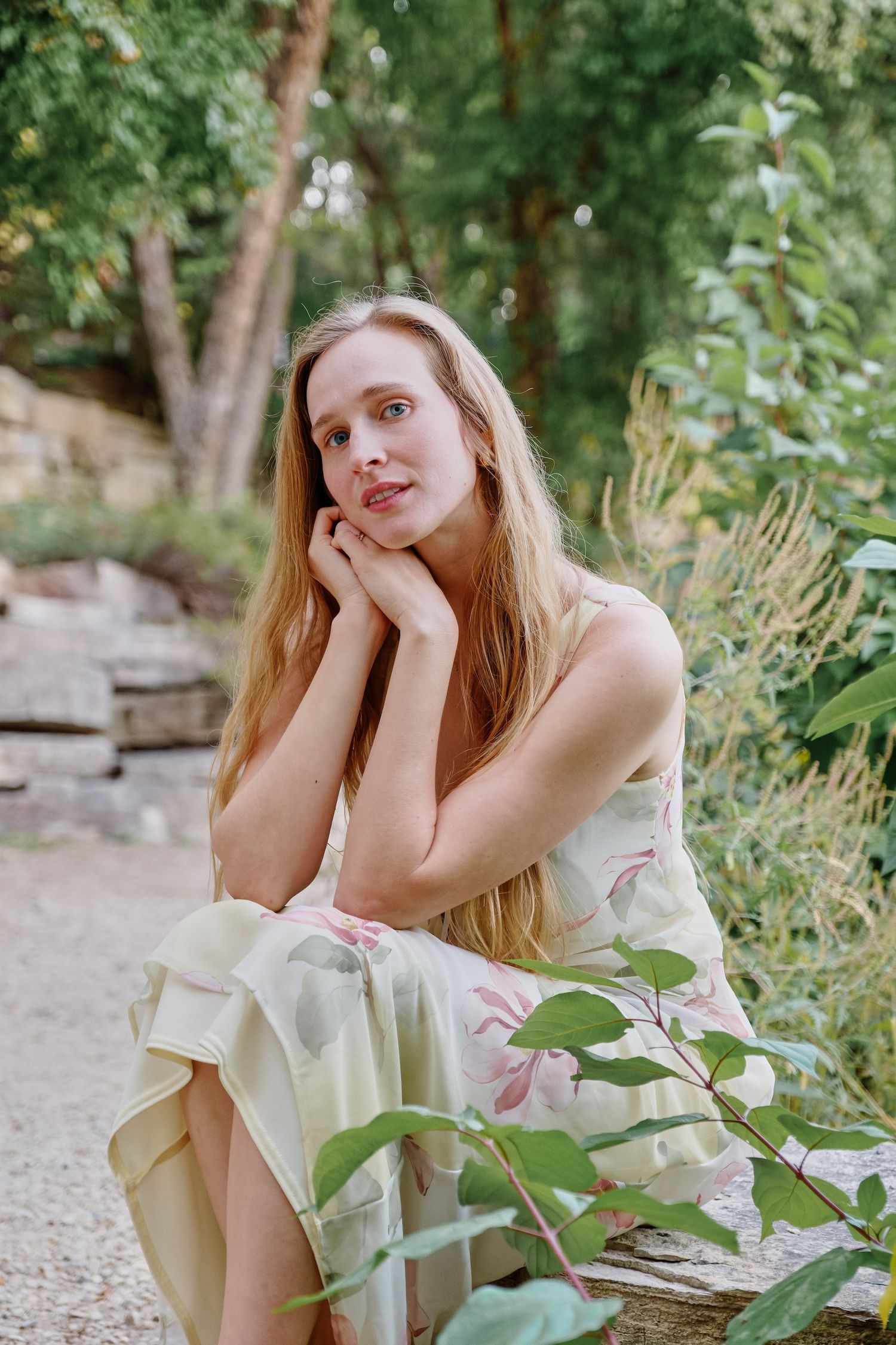 Young blonde woman in a floral dress sits outdoors surrounded by lush green garden foliage.