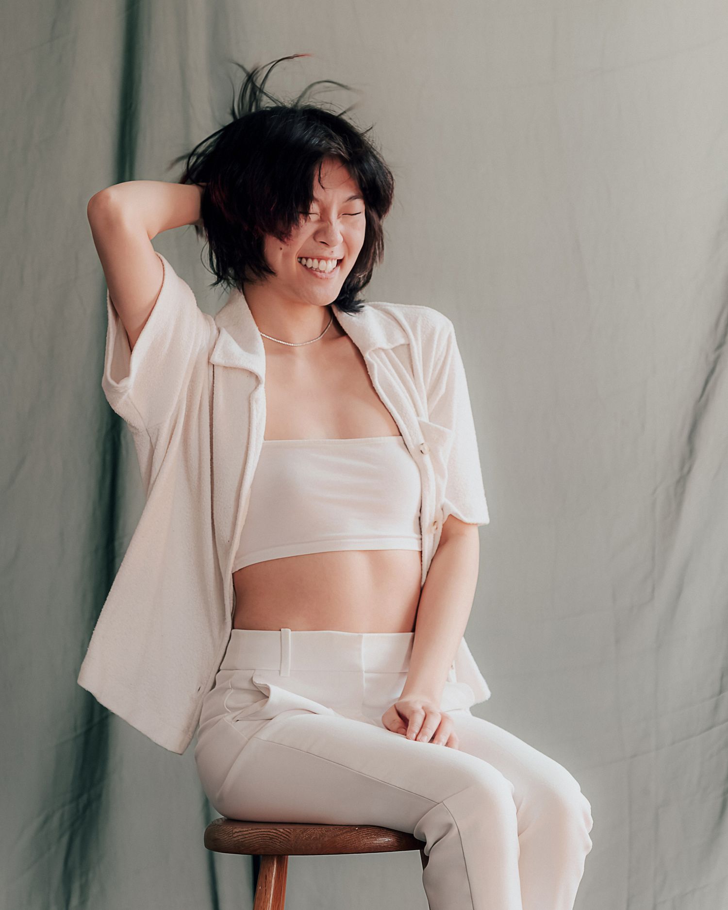 A smiling woman with short dark hair poses in white outfit on stool against gray backdrop.