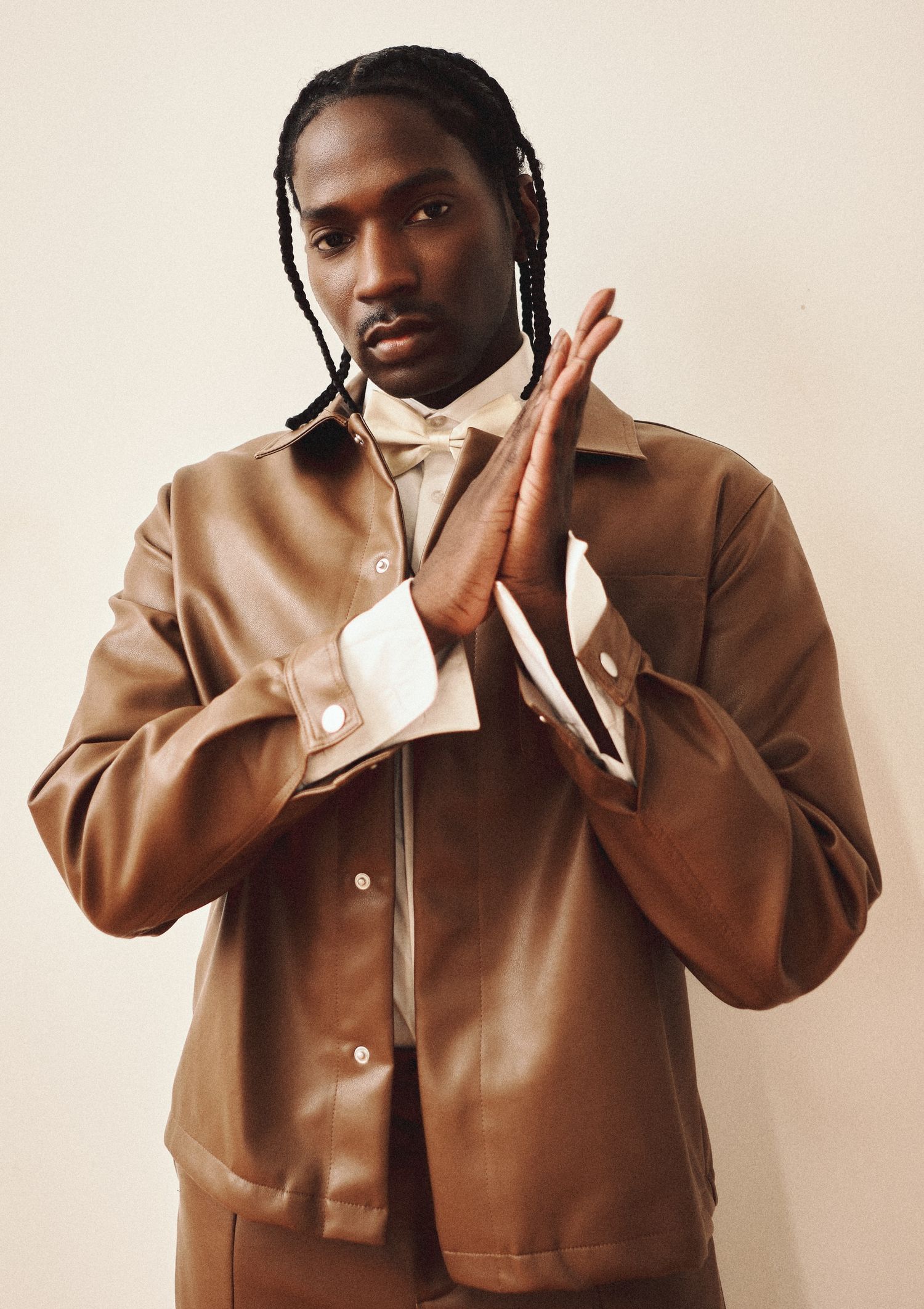 A man with braids wears a brown leather jacket and matching trousers, hands pressed together in a studio portrait.