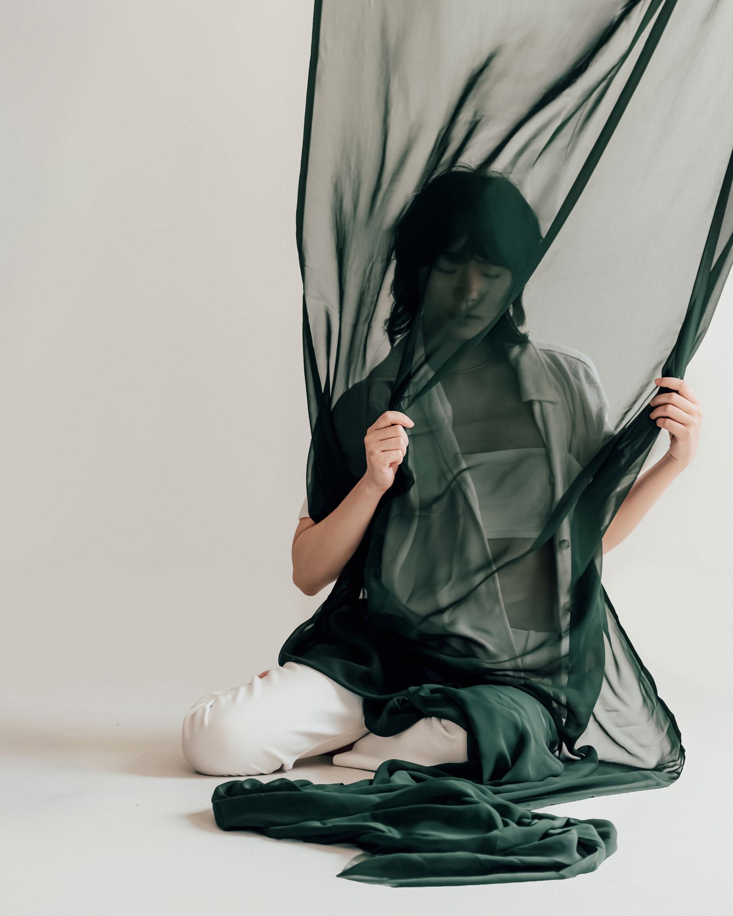 Person sitting on floor draped in flowing dark green sheer fabric against a light background.