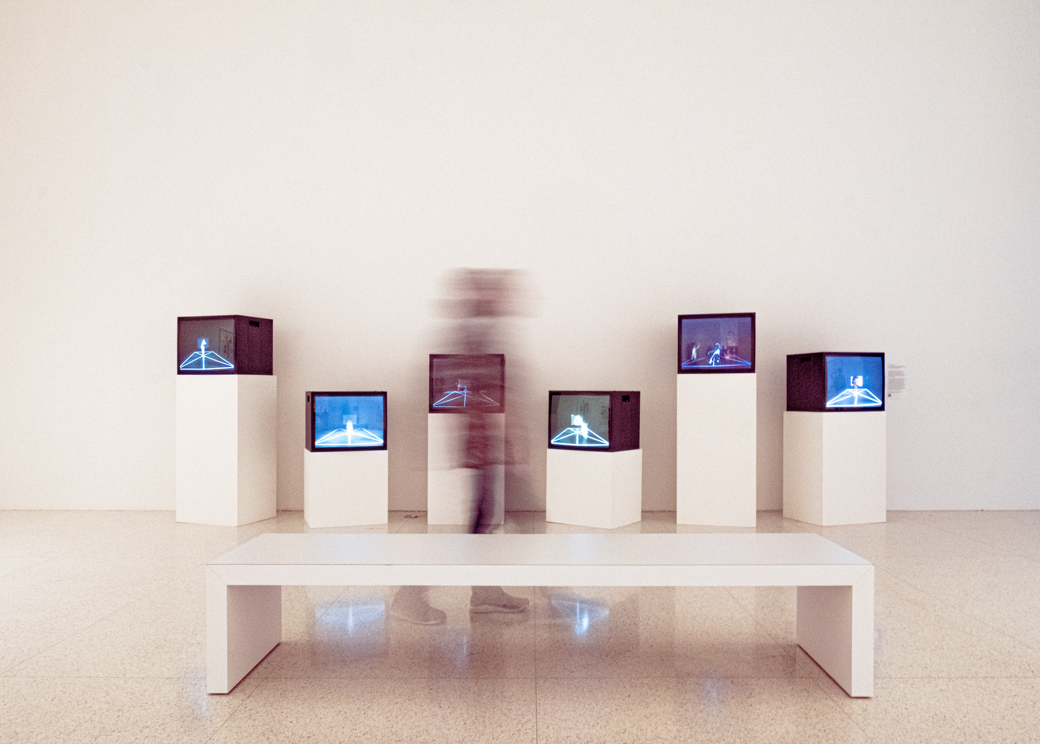 Blurred figure walks past multiple video art installations on white pedestals in a modern gallery space.