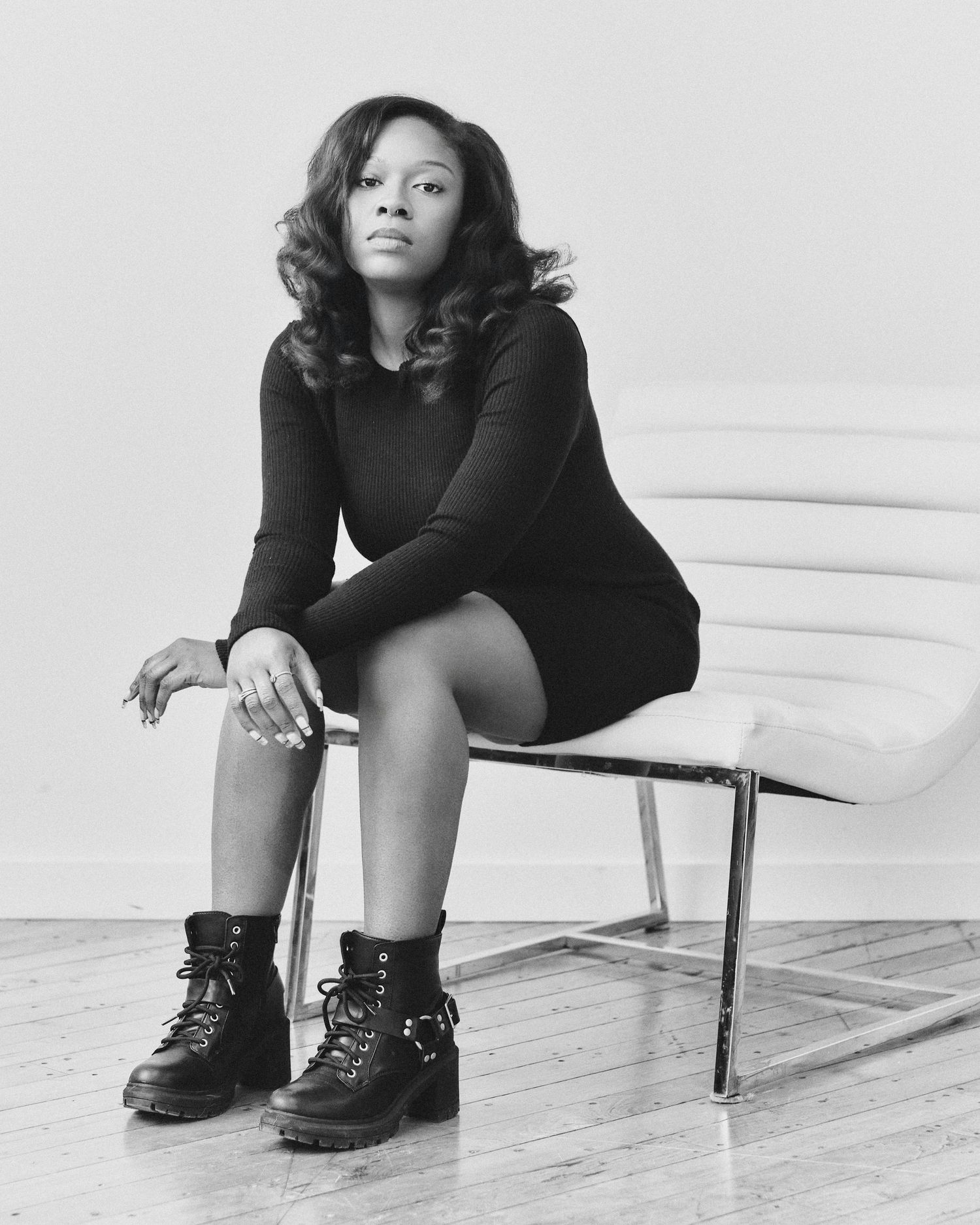 Black and white portrait of a young woman in a black dress and boots sitting on a white chair.