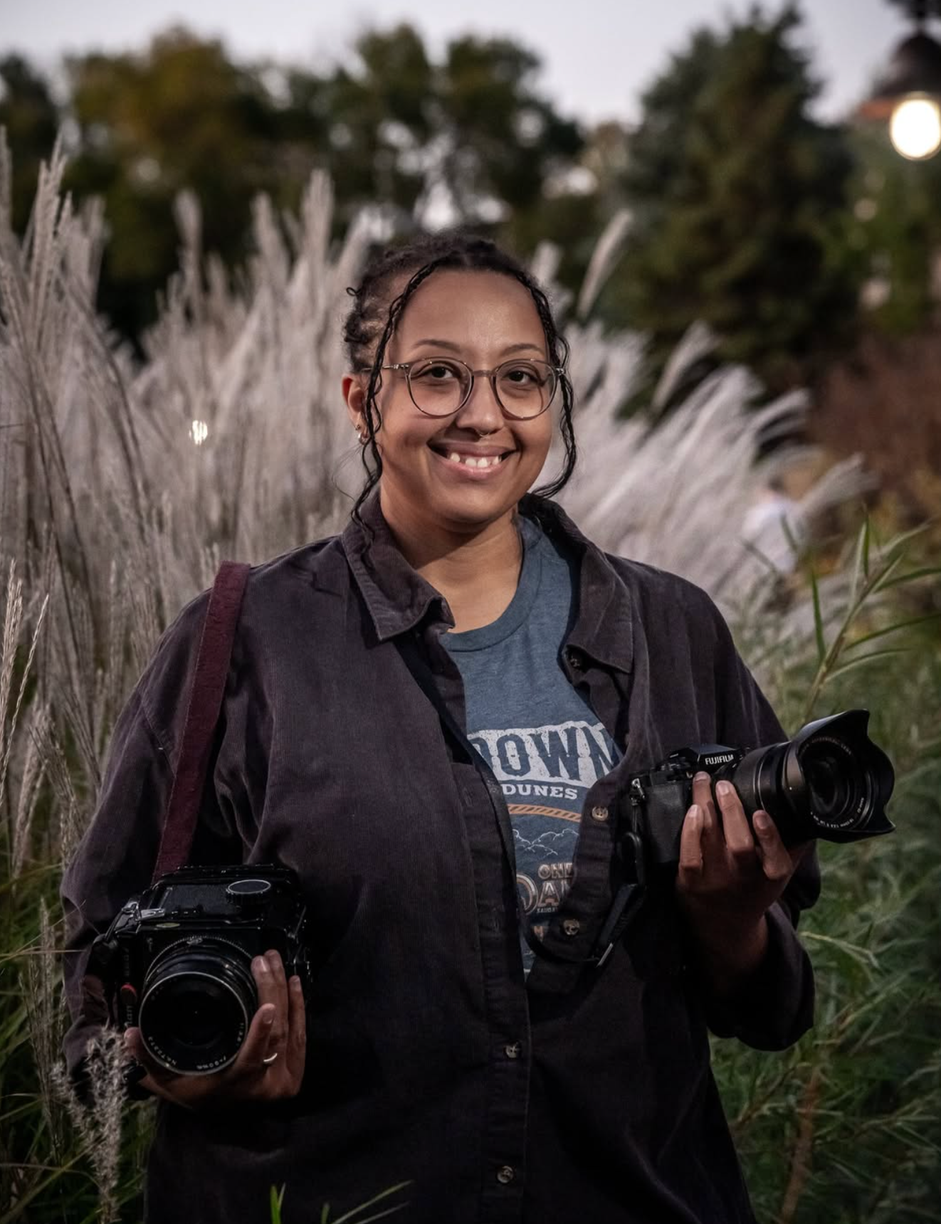 Smiling female photographer holding two cameras outdoors among ornamental grasses at dusk.