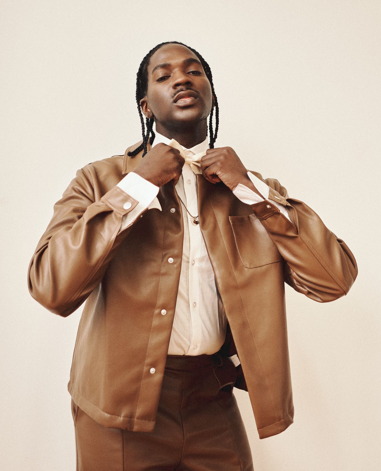 A man with braids adjusts his collar wearing an open brown leather jacket over a white dress shirt in a studio.