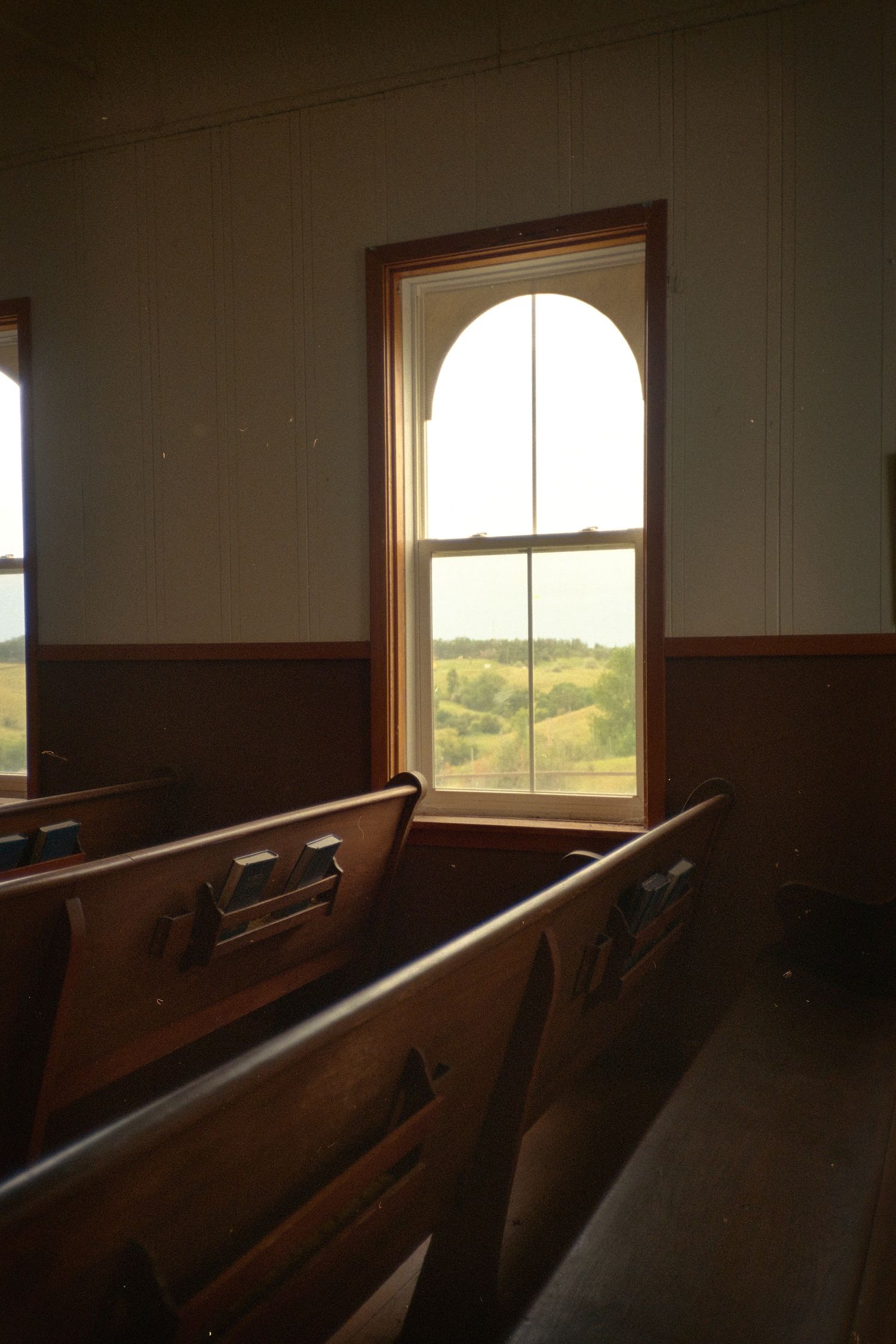 Wooden church pews beside an arched window overlooking a scenic green landscape.