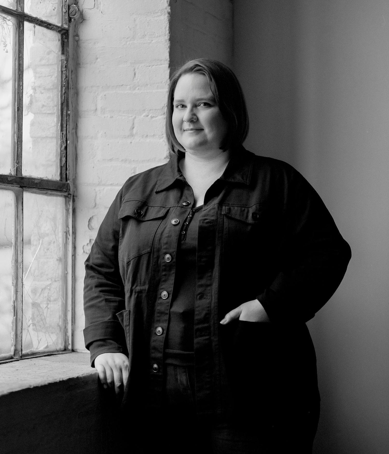 Black and white portrait of a woman in a denim jacket leaning against a brick wall by a window.