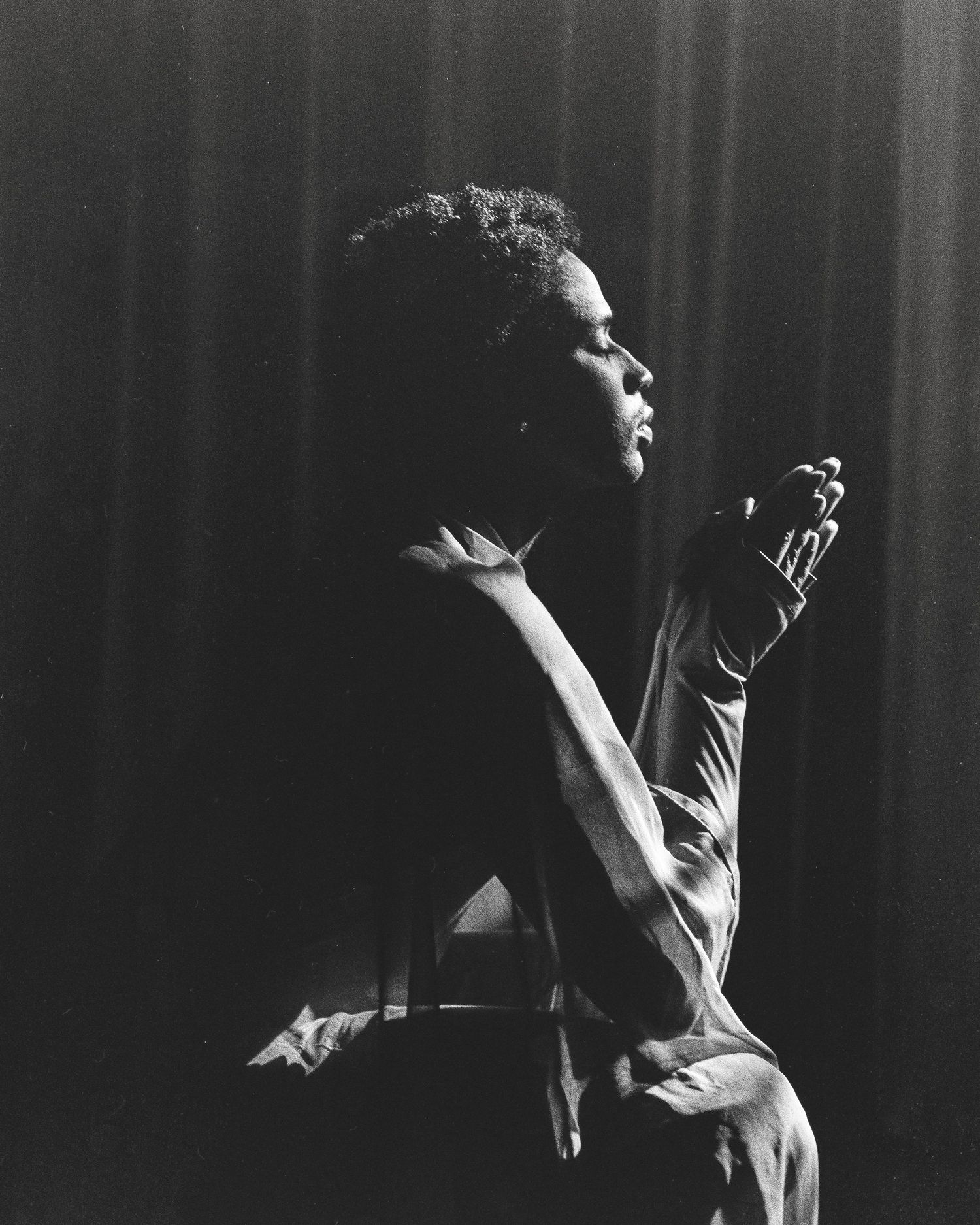 Black and white photo of a person sitting on stage, gazing upward with hand raised, dramatic curtain backdrop.