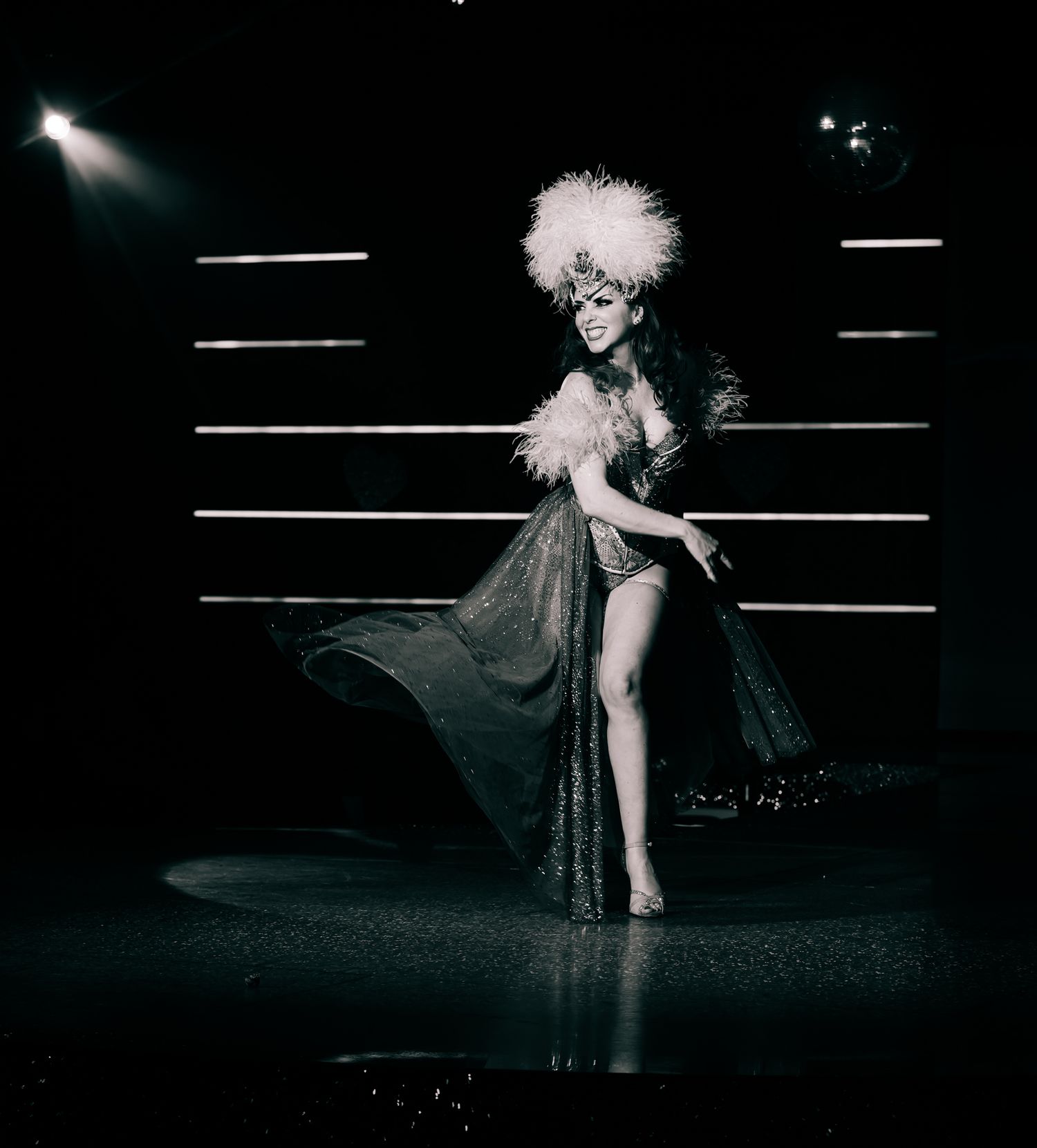 Elegant performer in feathered headdress and sparkling gown poses dramatically on a dark cabaret stage.