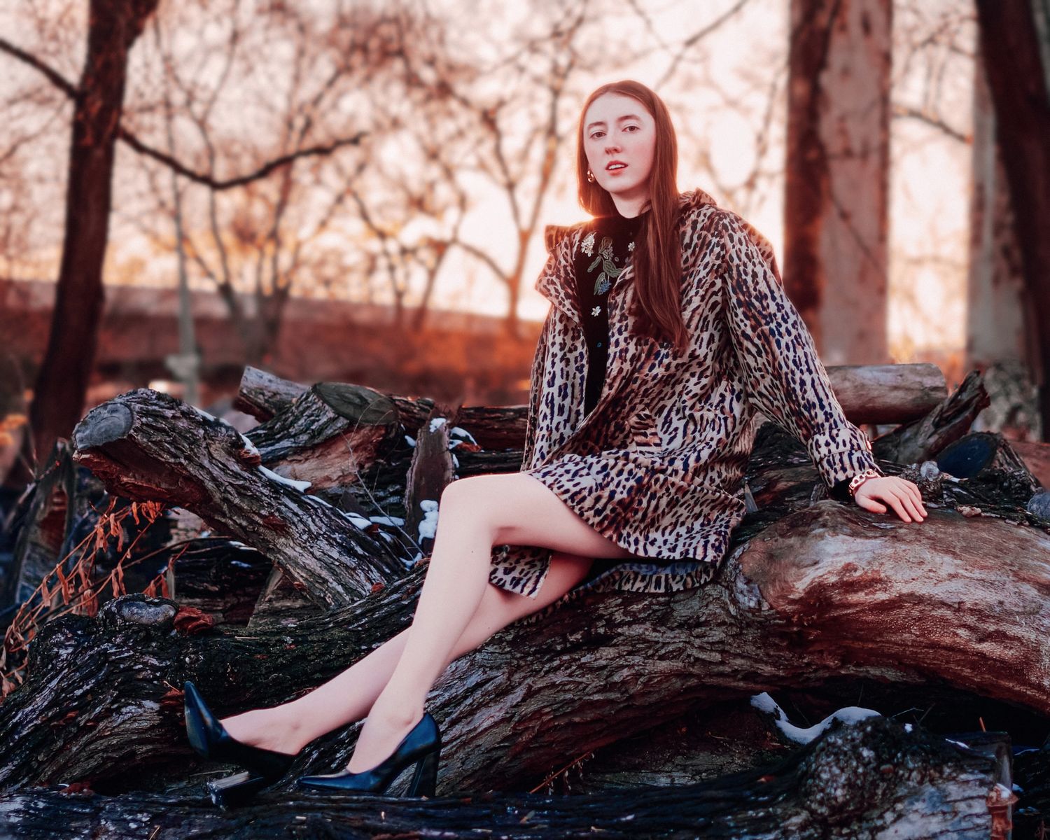 Redhead woman in leopard print coat sits on fallen logs during a golden sunset in a wooded area.