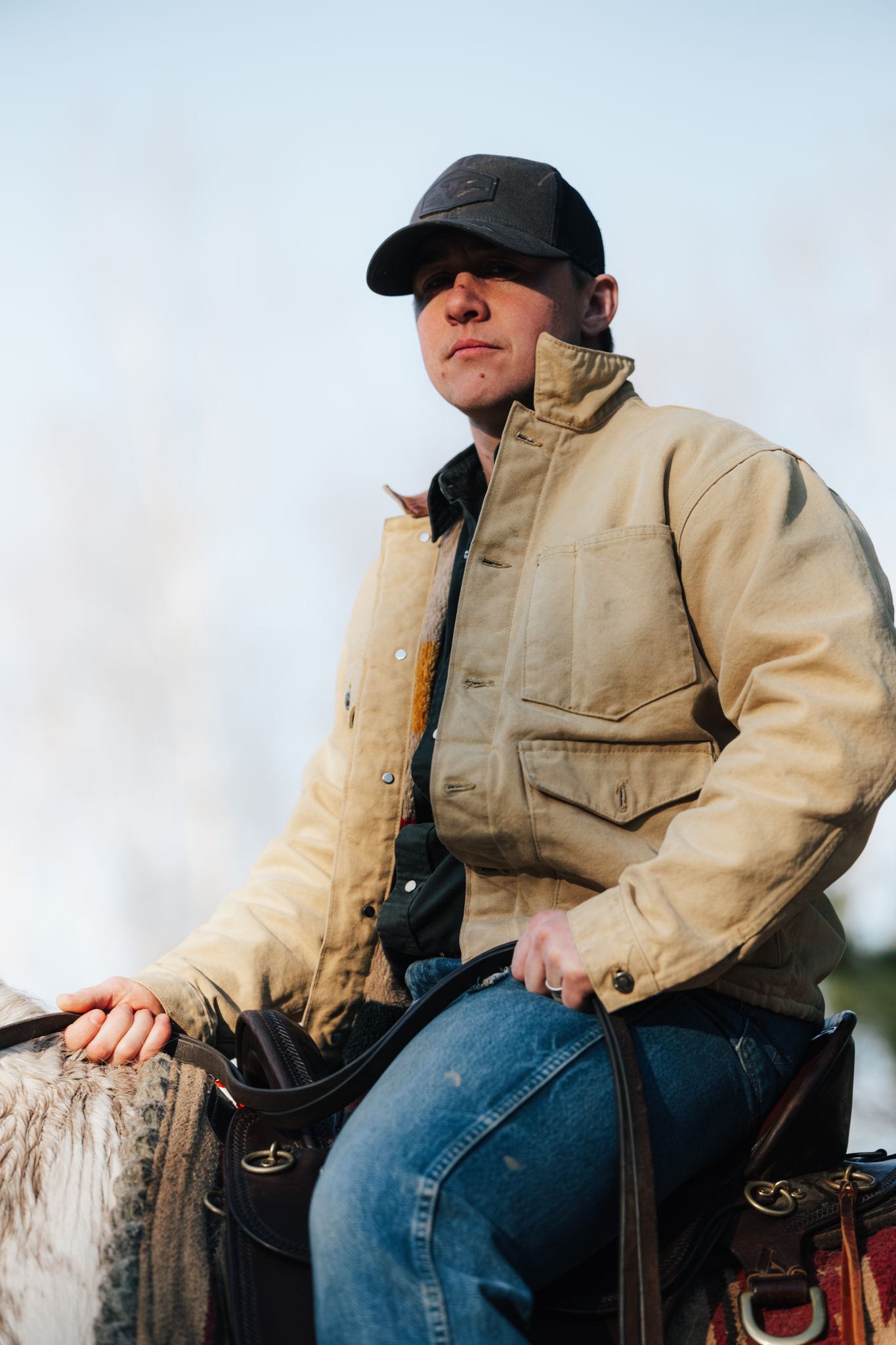 A man in a tan jacket and black cap rides a white horse outdoors in natural light.