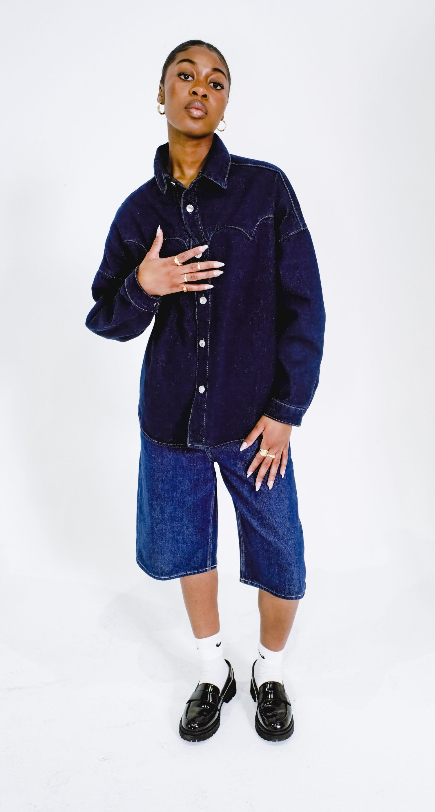 A woman poses in an oversized dark denim shirt, blue denim shorts, white socks, and black loafers on white background.