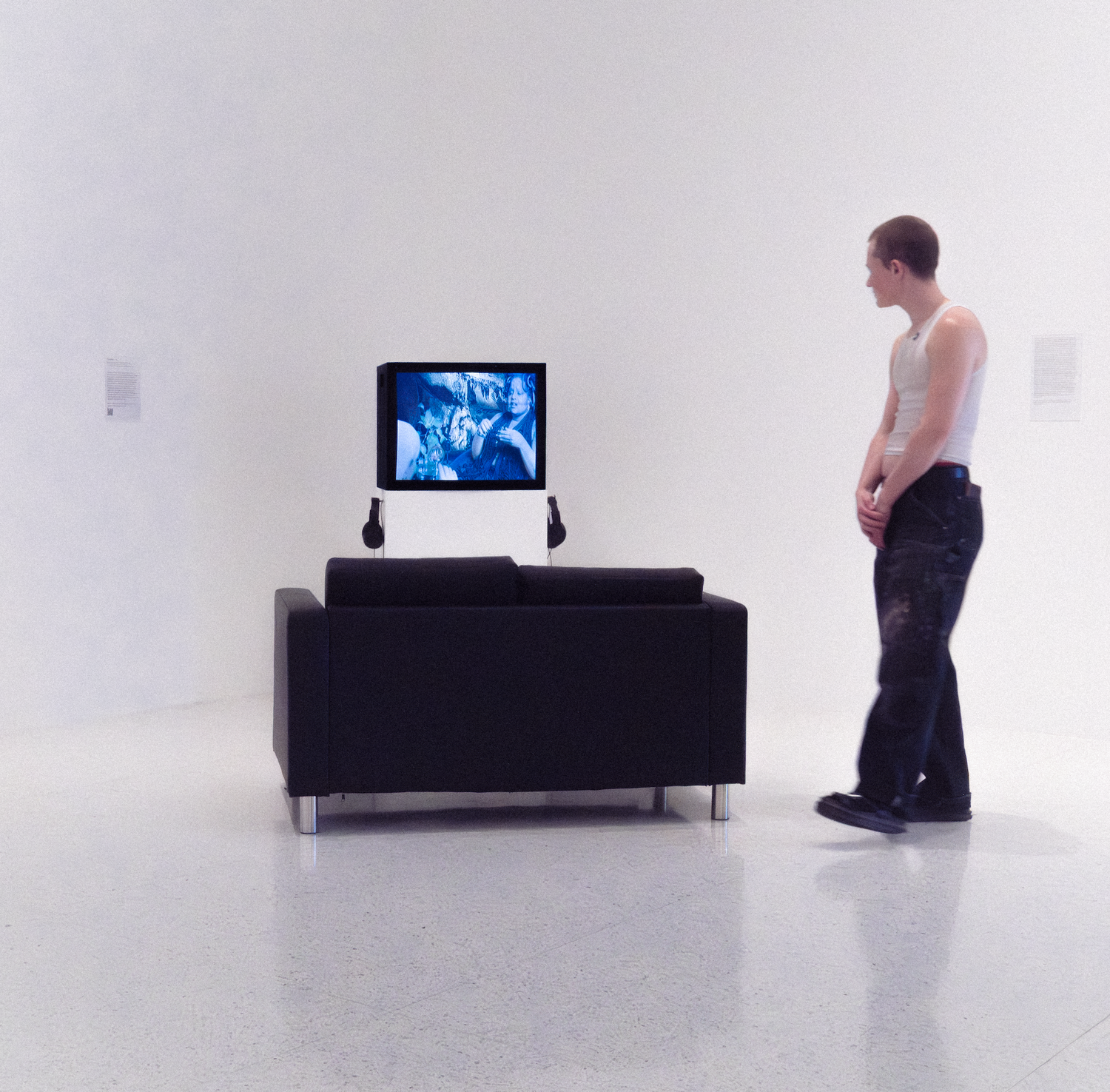Man standing in white gallery viewing a blue video artwork on a black sofa.
