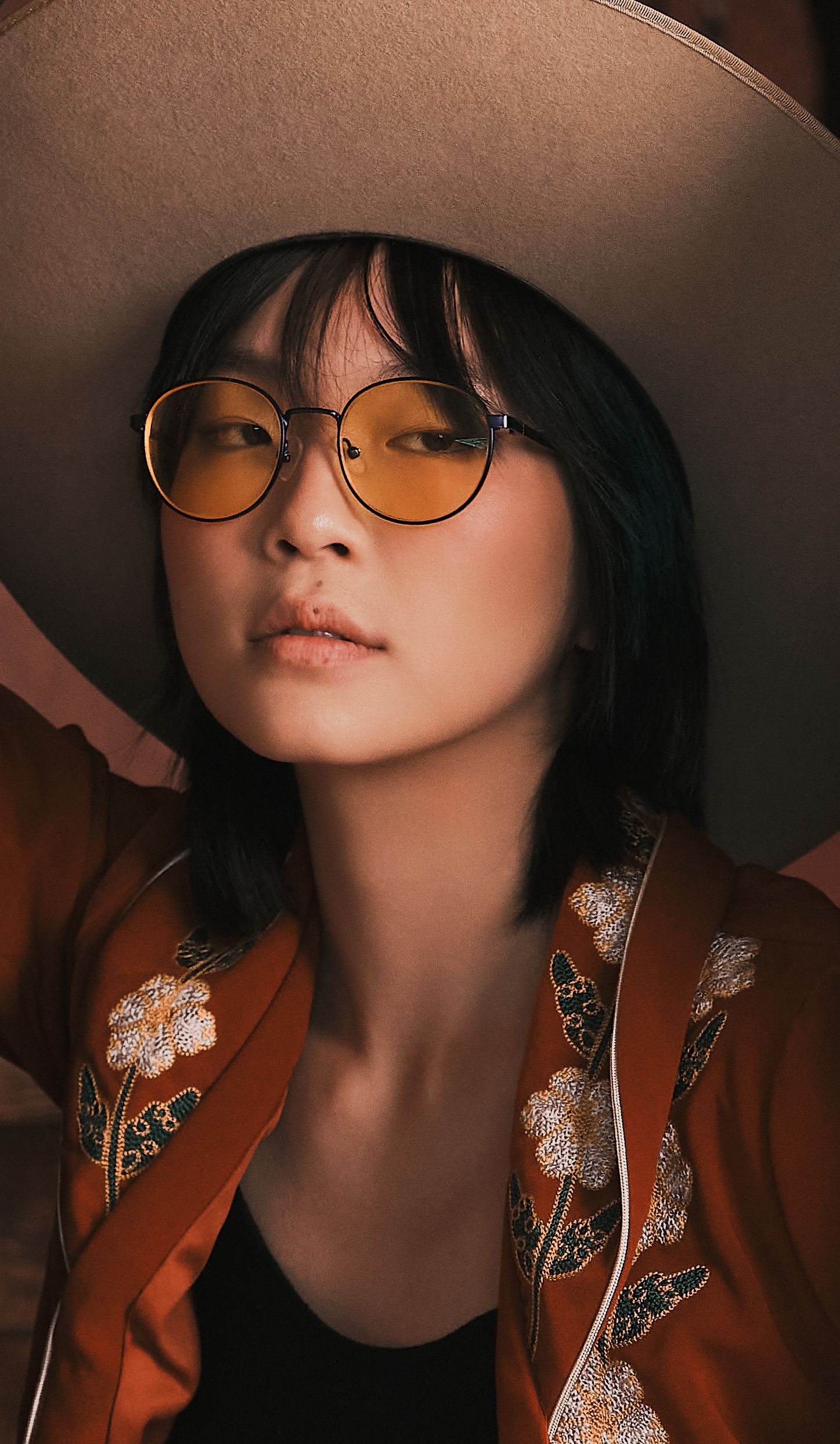 Asian woman wearing round yellow-tinted glasses, wide-brim hat, and embroidered orange jacket in warm light.