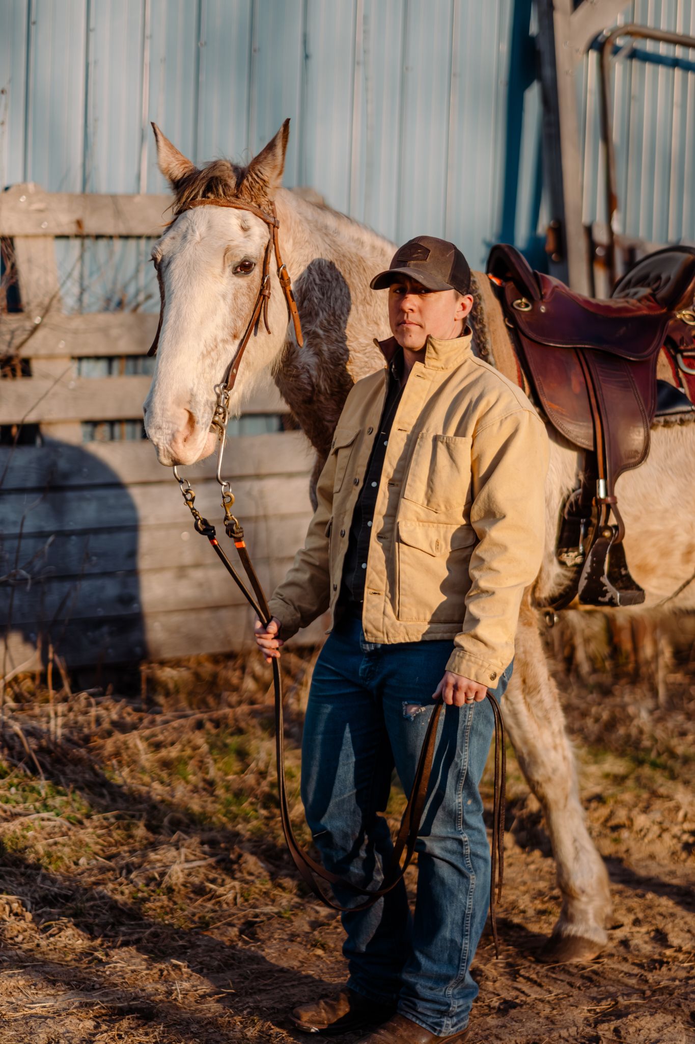 A cowboy standing beside a white horse near a stable in golden hour light.