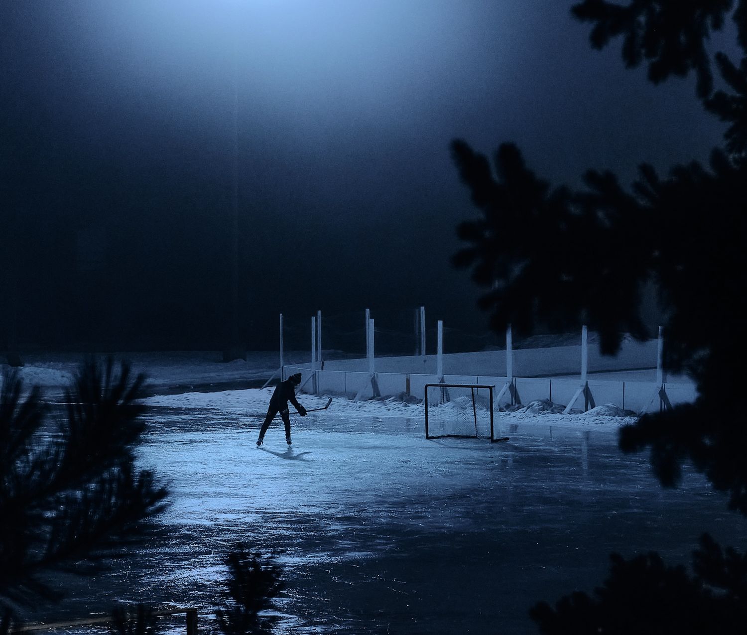 Lone hockey player skating on an outdoor rink under dramatic blue moonlit sky.