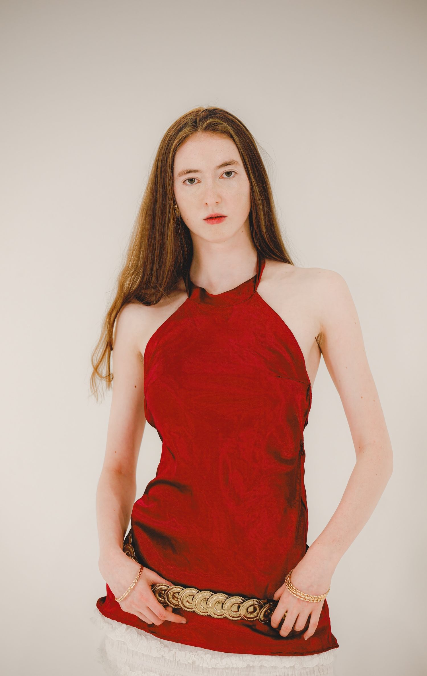 Young woman in red halter top with gold coin belt posing against white background.