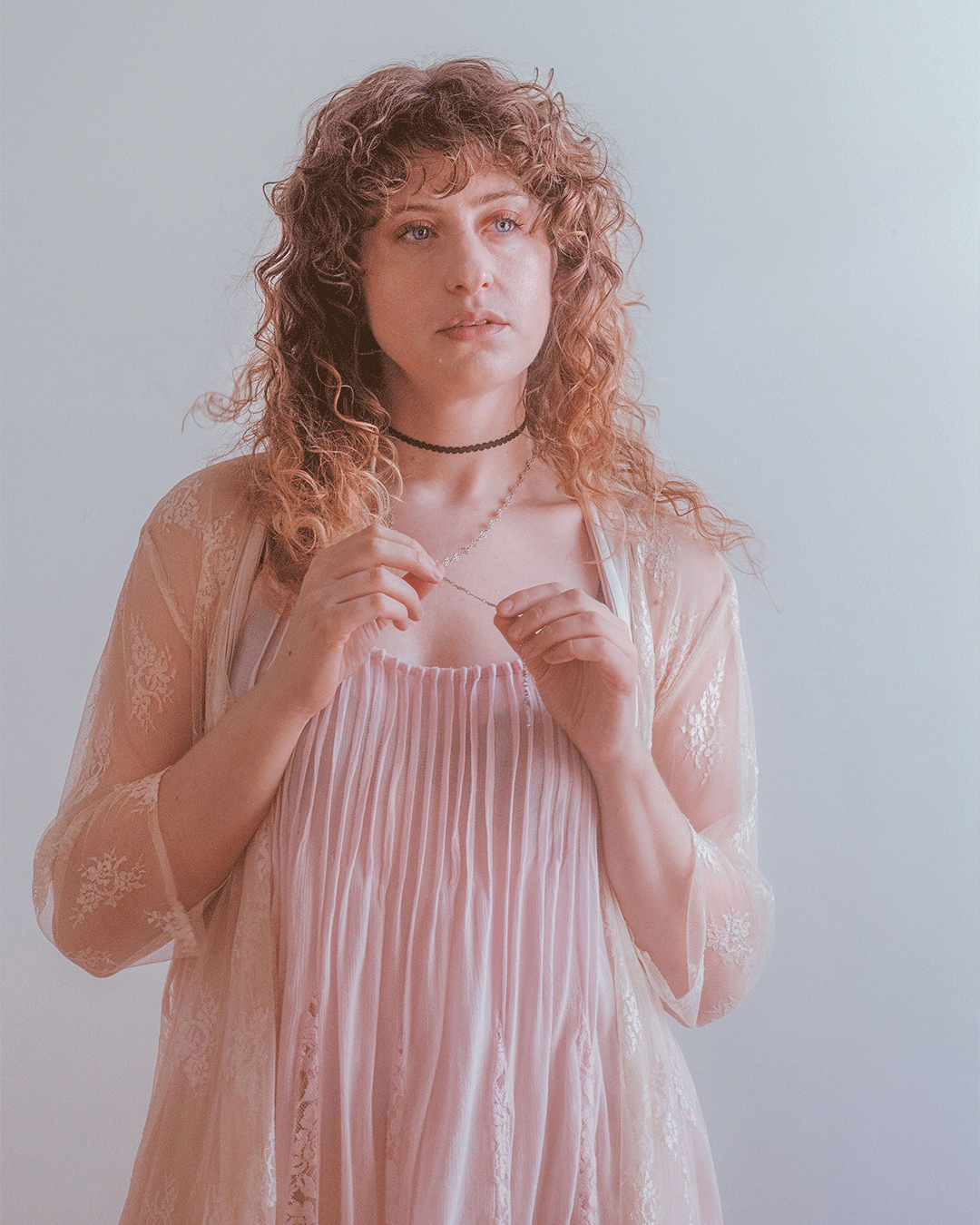 Young woman with curly auburn hair wearing a pink dress and lace kimono, holding a delicate necklace.