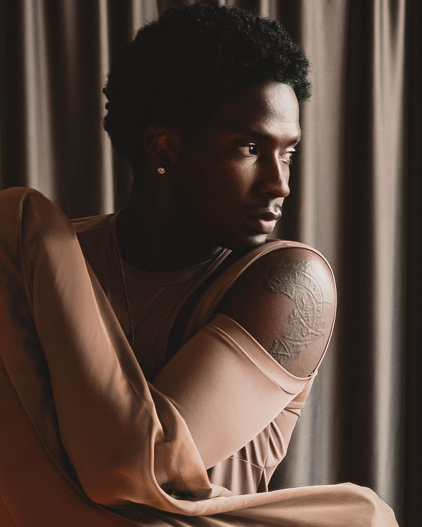 Young Black man with tattoo on shoulder wearing tan outfit, seated pensively against draped curtain backdrop.