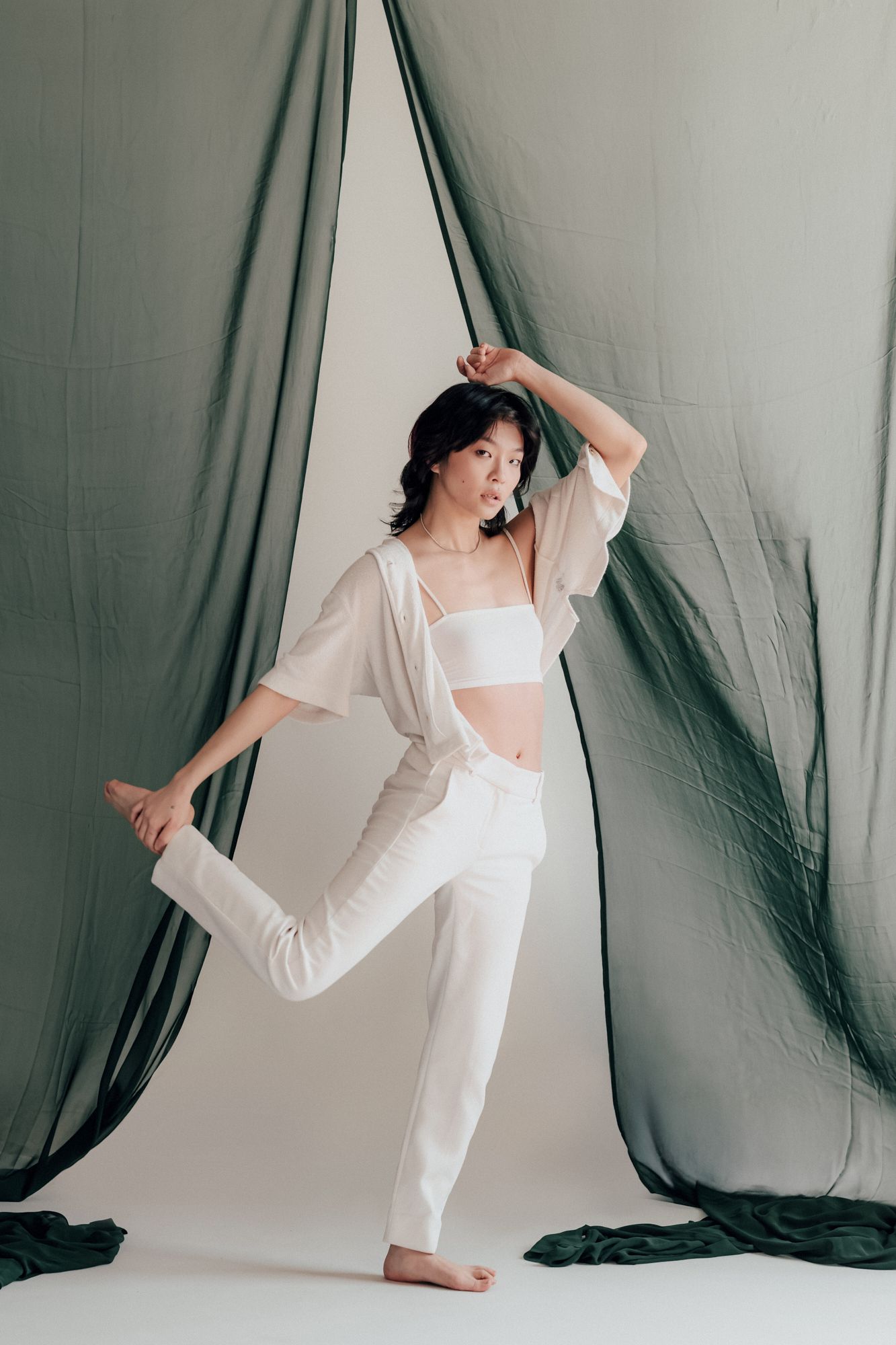 Woman in white crop top and pants poses gracefully barefoot against draped green fabric backdrop.