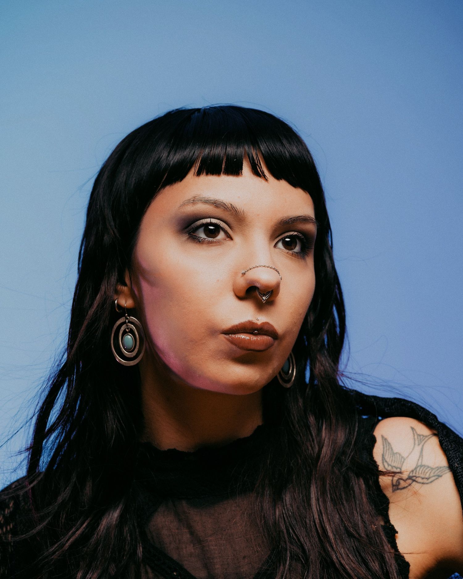 Woman with black hair, bangs, nose ring, and tattoo wears dark top against a blue background.