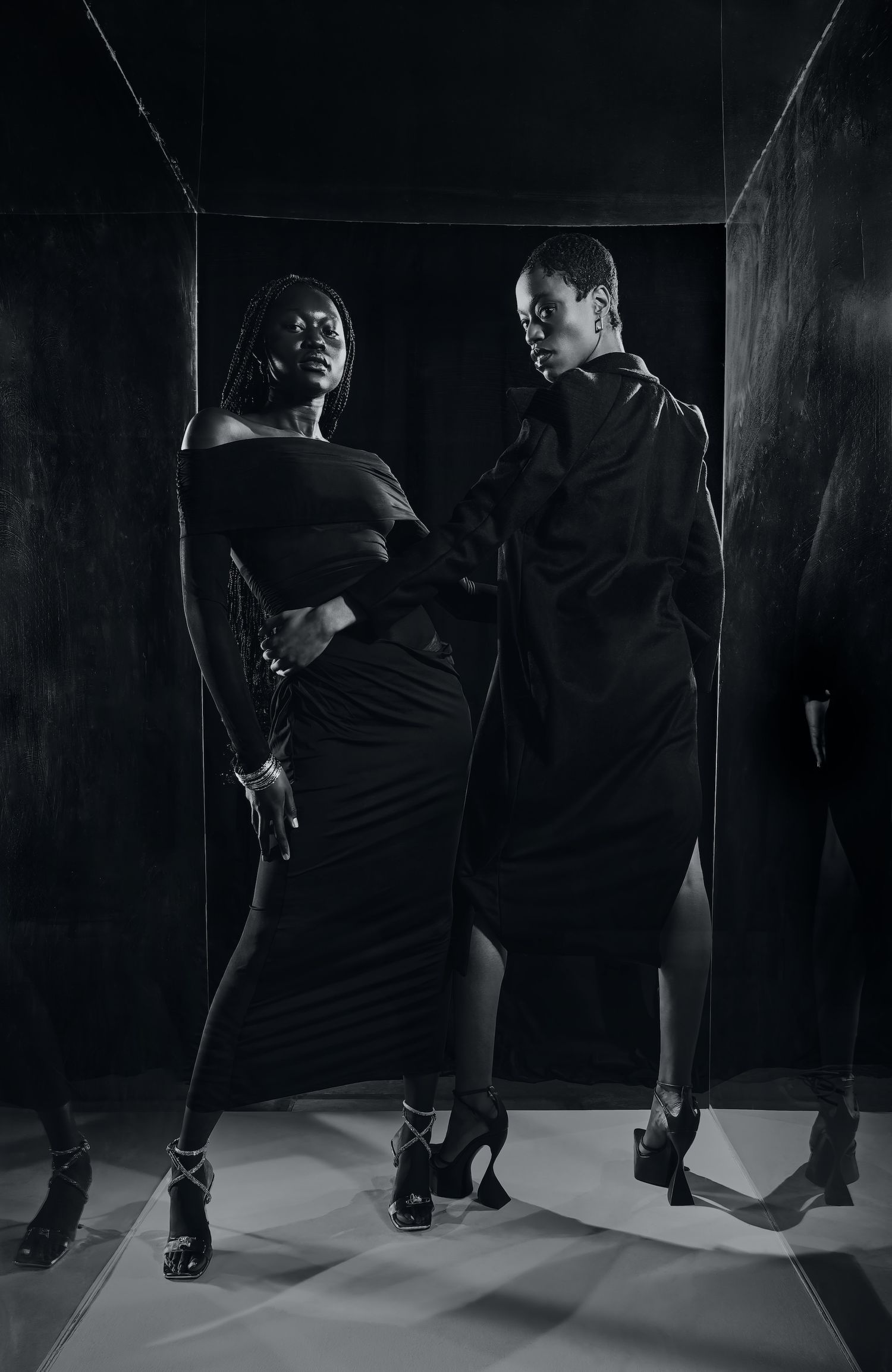 Two models in black outfits posing dramatically in dark studio setting, black and white photo.