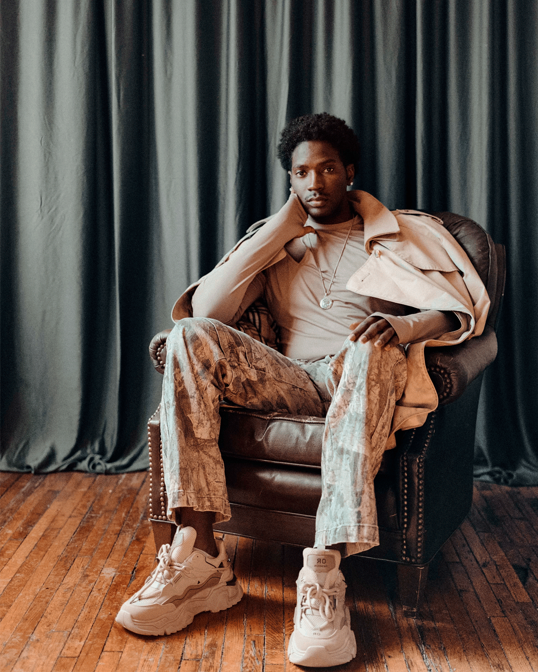 A stylish young Black man sits in a leather chair wearing a beige trench coat, patterned pants, and chunky sneakers.