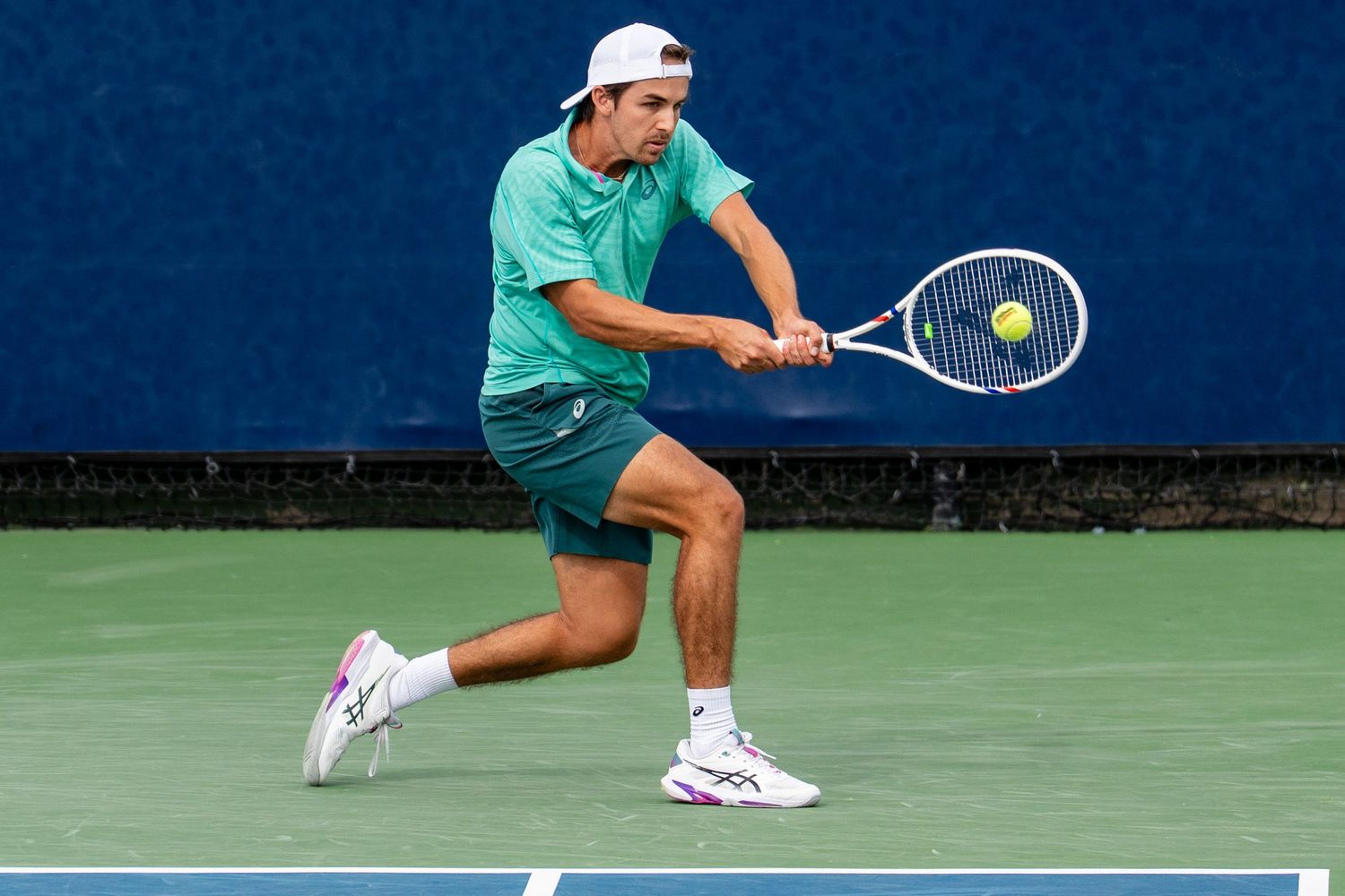 Male tennis player in green outfit hits backhand on blue hard court at San Diego Open.