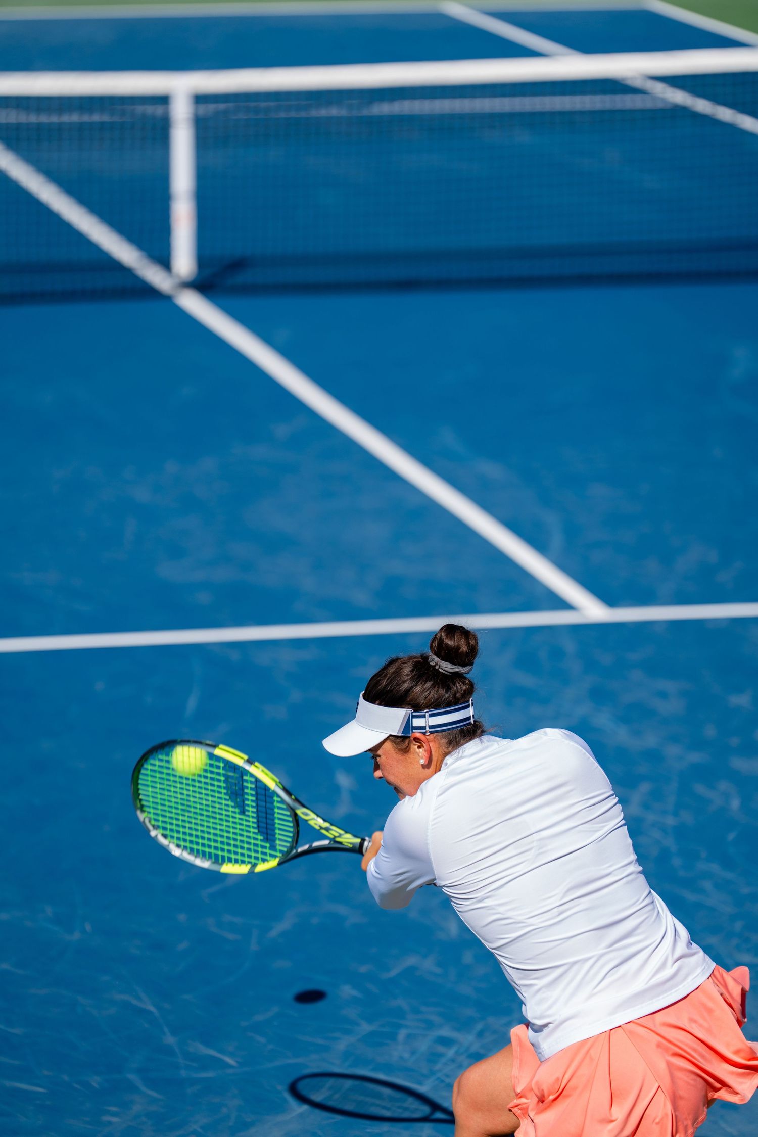 Female tennis player in white outfit prepares backhand shot on blue hard court from behind.