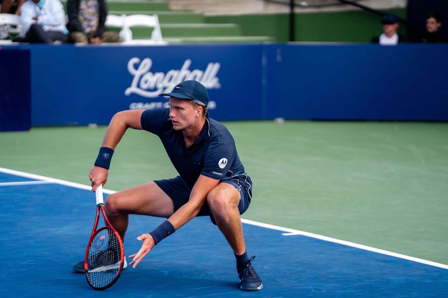 Tennis player crouches low for backhand return on blue Longball hard court.