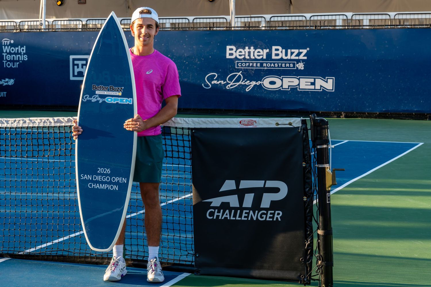 Tennis player poses with trophy and surfboard at ATP Challenger San Diego Open on blue hard court.