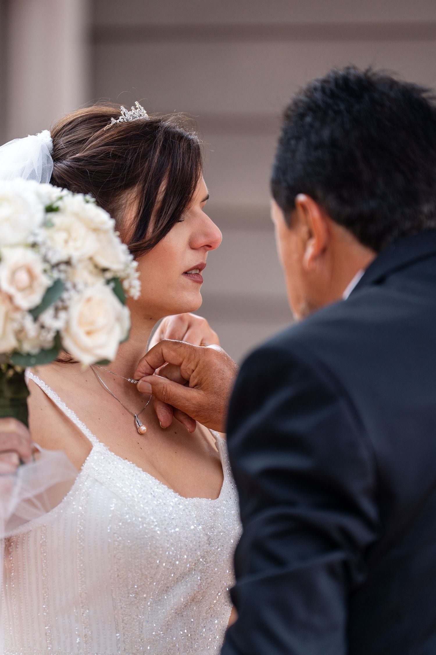 Groom fastening a necklace on bride in white sequin dress and tiara, holding bouquet of white roses.