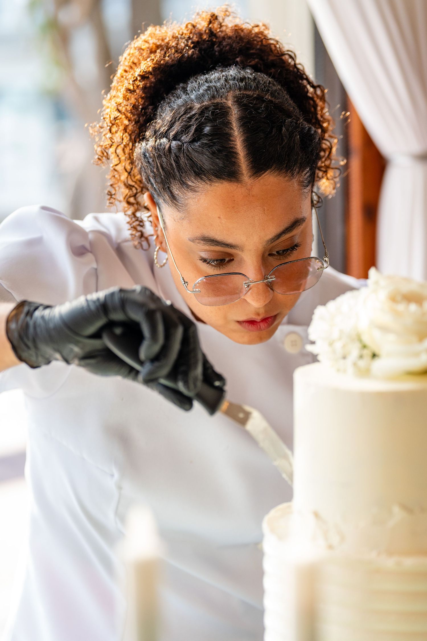 A pastry chef with curly hair and glasses decorates a white tiered wedding cake using a spatula while wearing black gloves.
