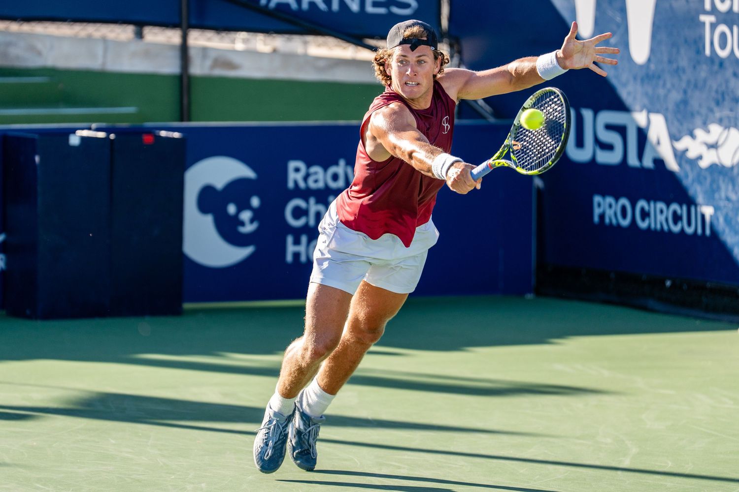 Male tennis player in red lunges to return a shot at a USTA Pro Circuit tournament.