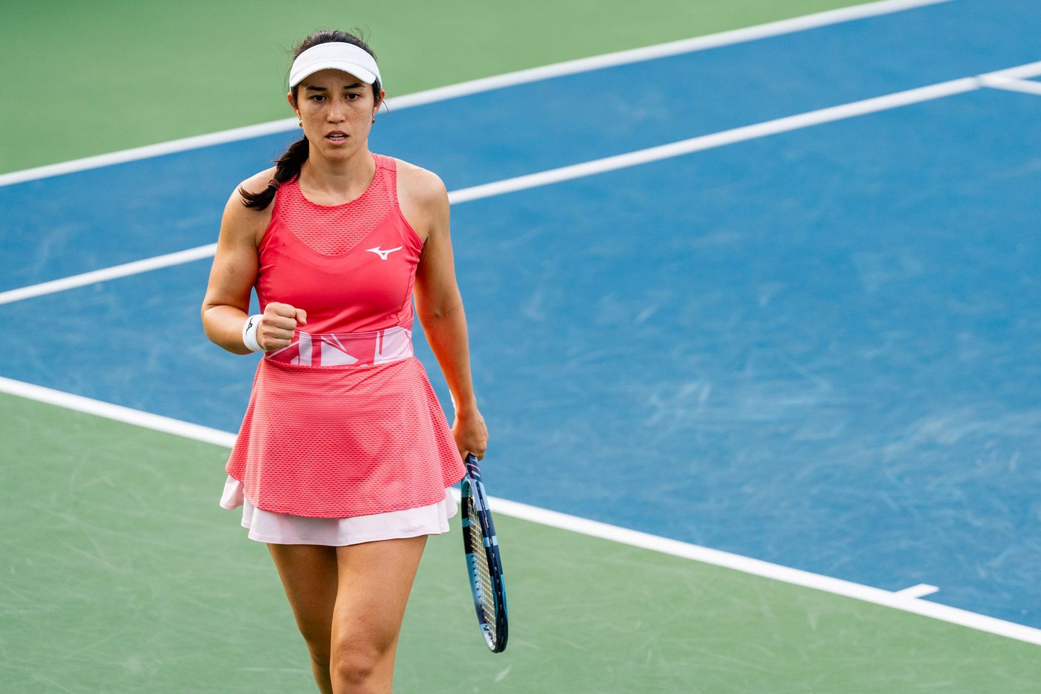 Female tennis player in pink Mizuno outfit holds racket on blue hard court.